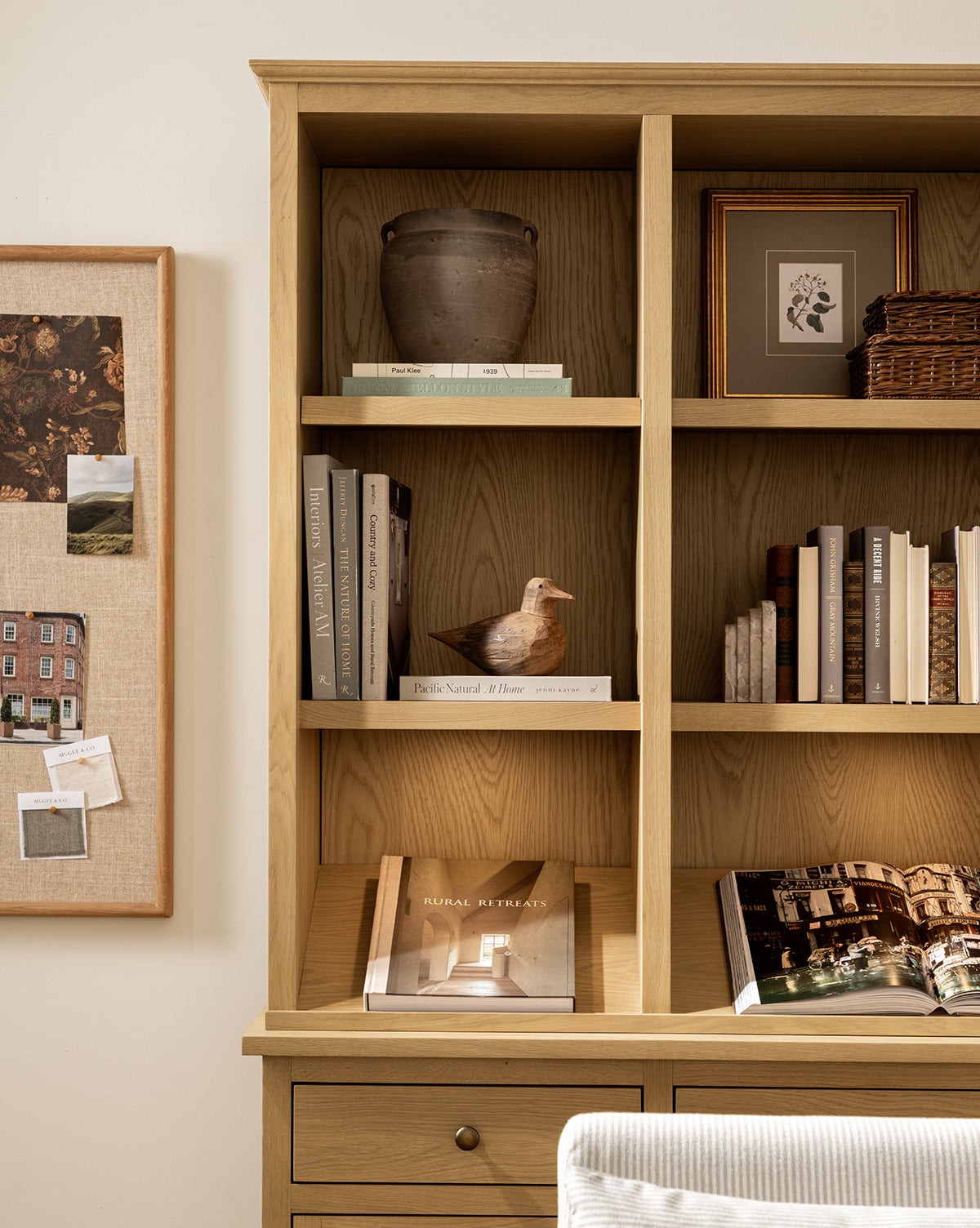 A wooden bookshelf displays books, a ceramic vase, a small framed artwork, a wicker basket, and the Wooden Bird Object to add nature-inspired charm. A bulletin board with photos and notes hangs on the wall to the left, by McGee & Co.