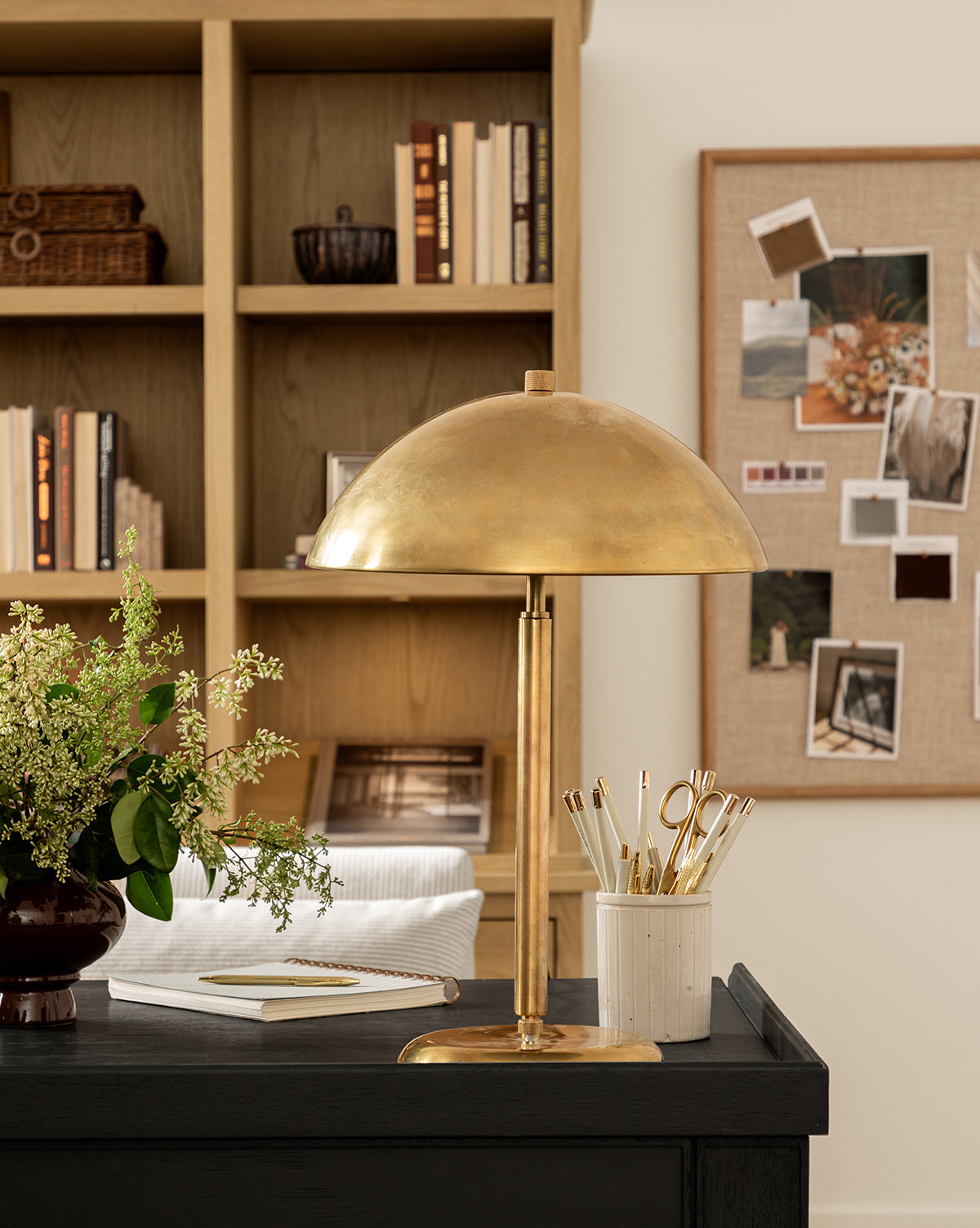 The Aldorno Desk Table Lamp sits on a black desk with gold pens, a plant, and a notebook. Behind it, wooden shelves hold books and baskets, while a bulletin board displays photos—perfect for stylish living room lighting, by McGee & Co.