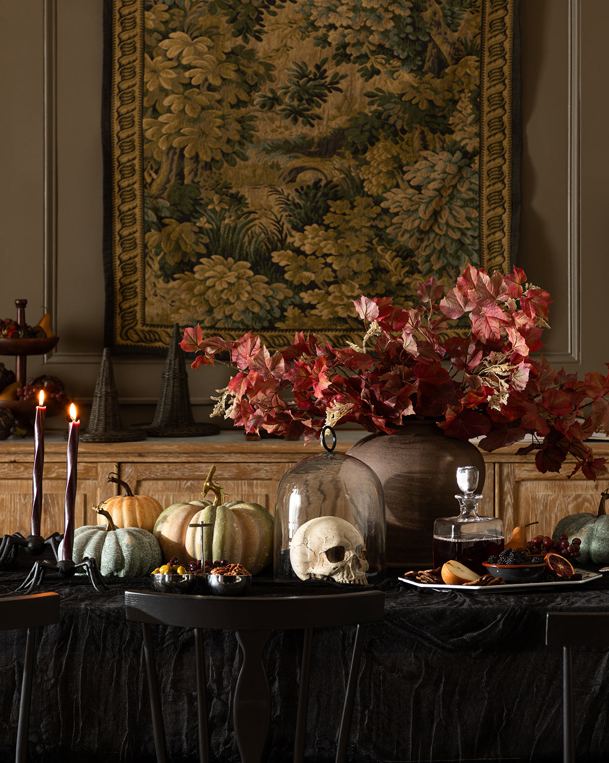 A moody Halloween table features pumpkins, a glass-domed skull, red leaves in a vase, tall black candles, and treats. The Hortensia Wall Tapestry with its leafy design hangs in the background for an elegant touch, by McGee & Co.