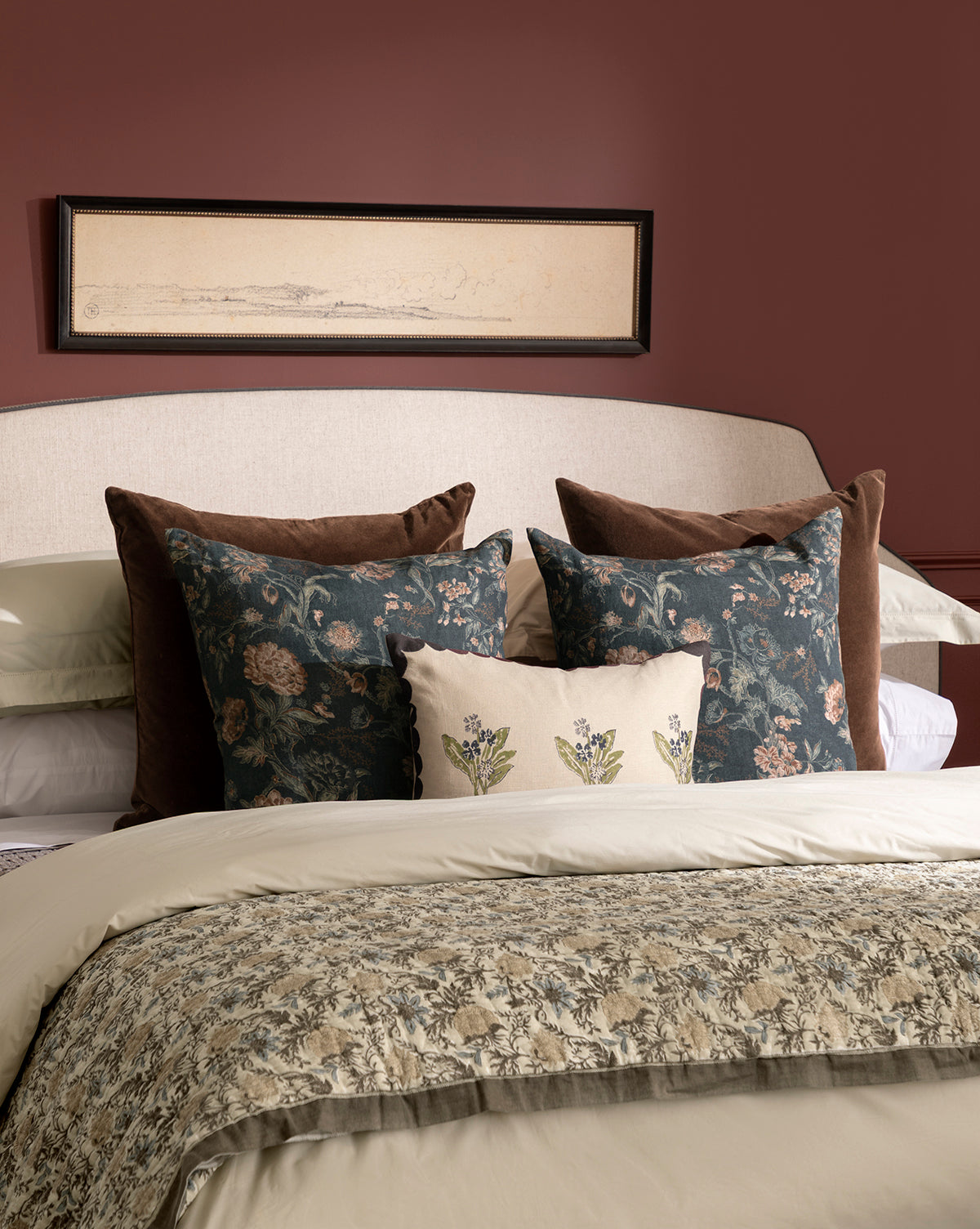 A neatly made bed dressed in Clouded Island bedding with cream and patterned blankets and layered floral embroidered pillows, set before a padded headboard and mauve wall featuring a black-framed landscape sketch above, by McGee & Co.