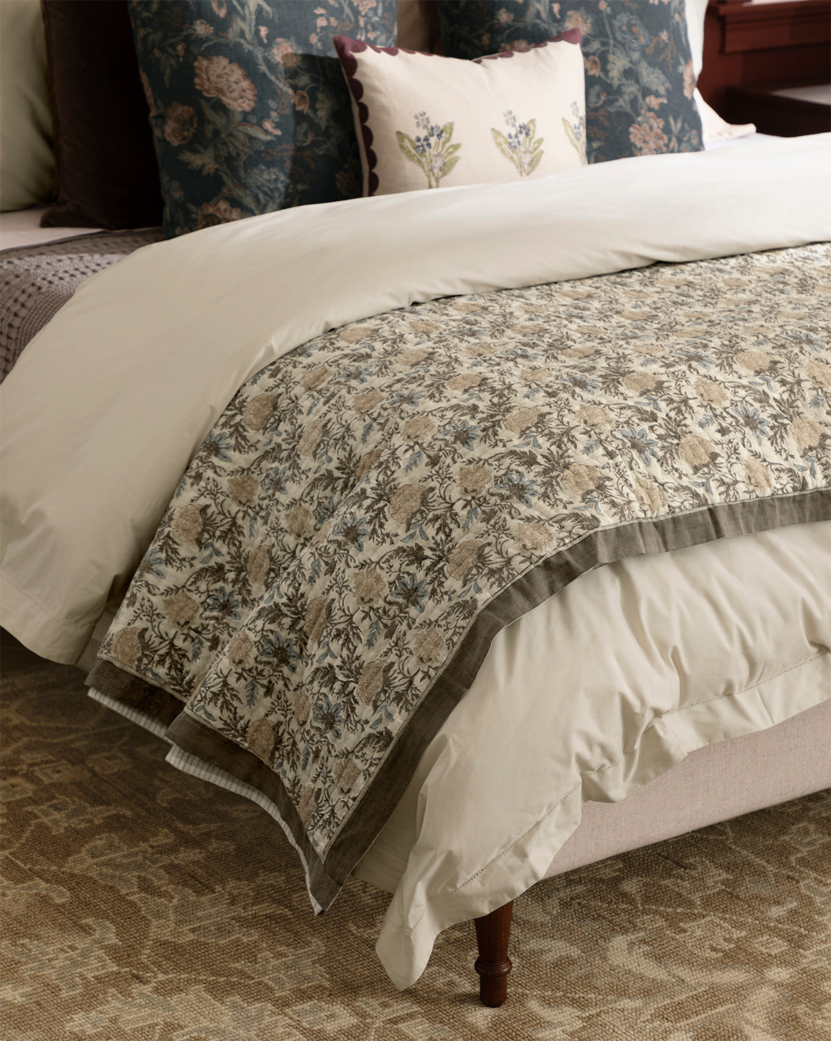 A cozy bedroom features a neatly made bed with cream-colored bedding, the Audrey Tan Block Print Quilt with ruffle edging, and floral decorative pillows, set atop a patterned carpet, by McGee & Co.