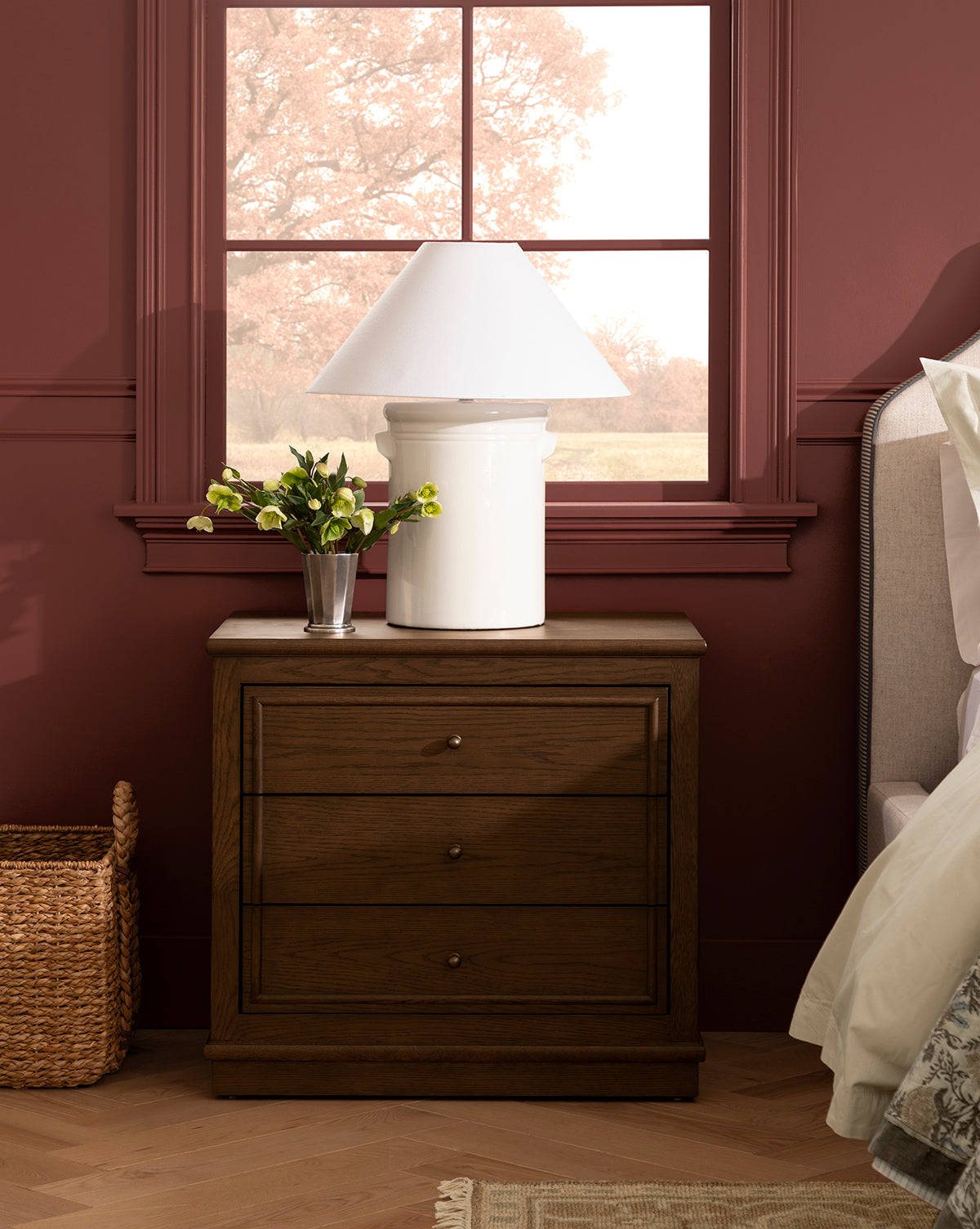 A Yorkshire Nightstand with an English oak finish and antique brass pulls stands by the bed, topped with a white lamp and vase of yellow flowers. Pinkish-red window trim frames leafy trees, and a wicker basket sits on the floor nearby, by McGee & Co.