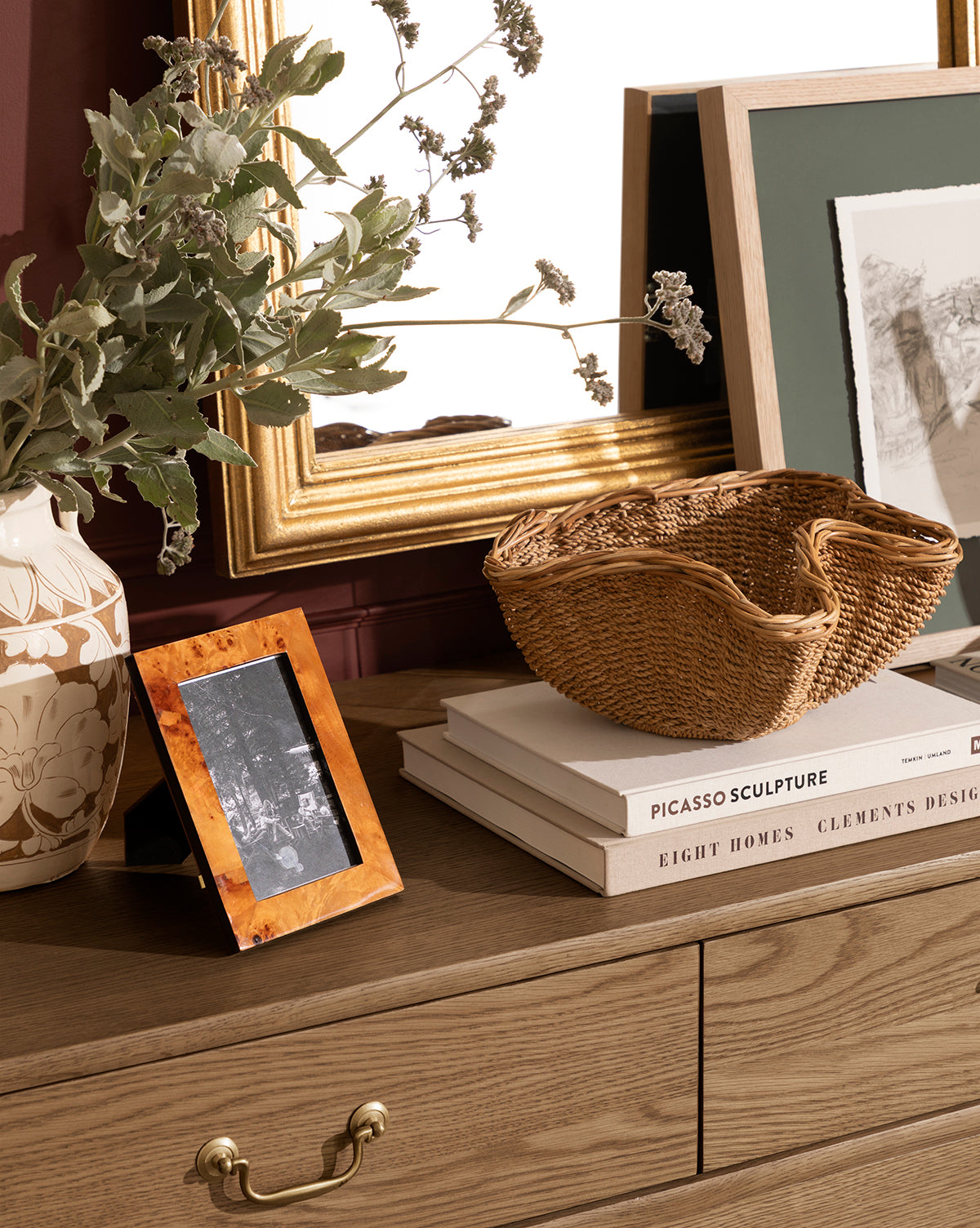 A wooden dresser with a gold-framed mirror, entryway decor like a ceramic vase with greenery, the Goulding Woven Bowl, a framed photo, and stacked books including one titled PICASSO SCULPTURE, by McGee & Co.