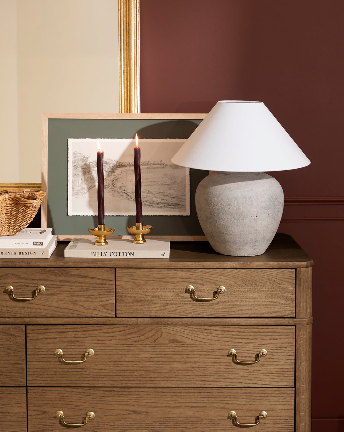 A Montague Table Lamp with a white shade sits atop a wooden dresser with brass handles, accompanied by two lit burgundy candles in gold holders, books, a wicker basket, and framed art against a maroon and gold-accented wall, by McGee & Co.
