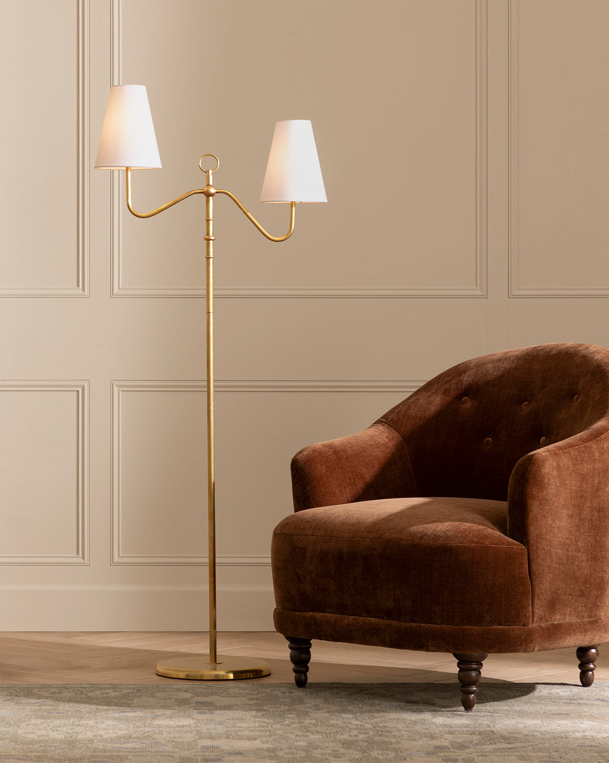The Makerspalm Holloway Floor Lamp, in antique brass with a double-arm and two white shades, stands next to a brown velvet armchair with wooden legs on a patterned rug beside a beige paneled wall, McGee & Co.
