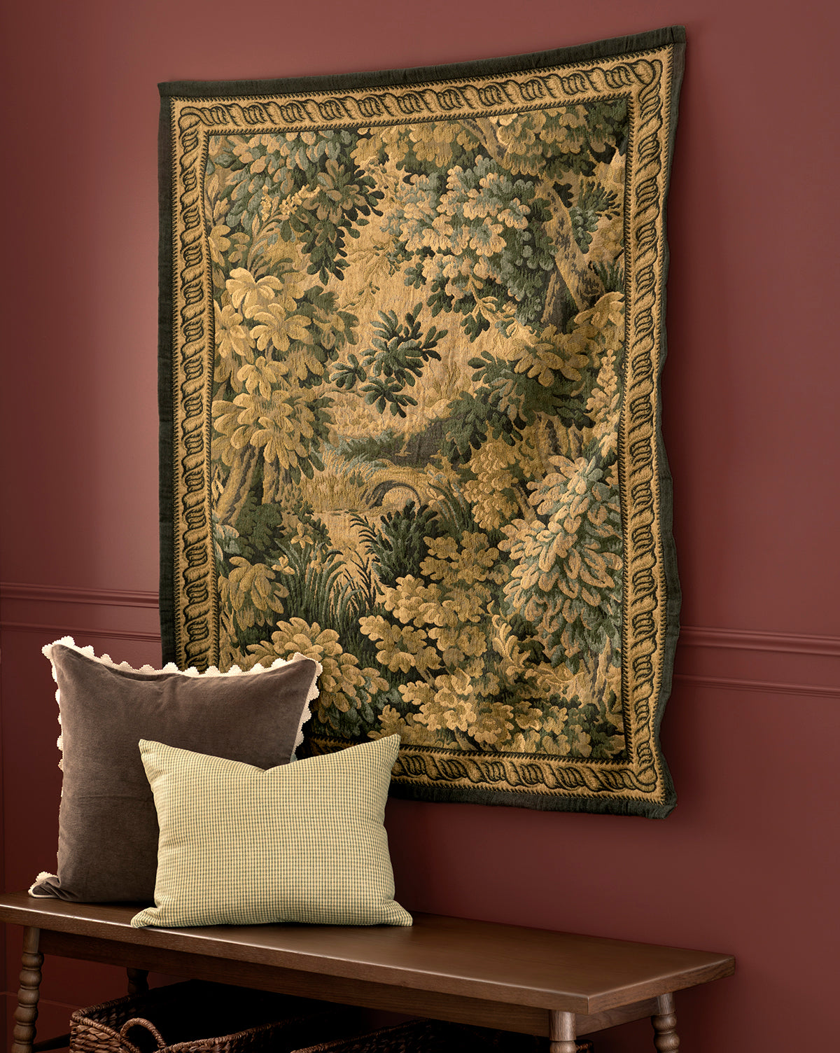 The Hortensia Wall Tapestry, featuring a leafy forest scene, hangs on a red wall above a wooden bench with two throw pillows: one brown with a scalloped edge and one light green with a checkered pattern, by McGee & Co.