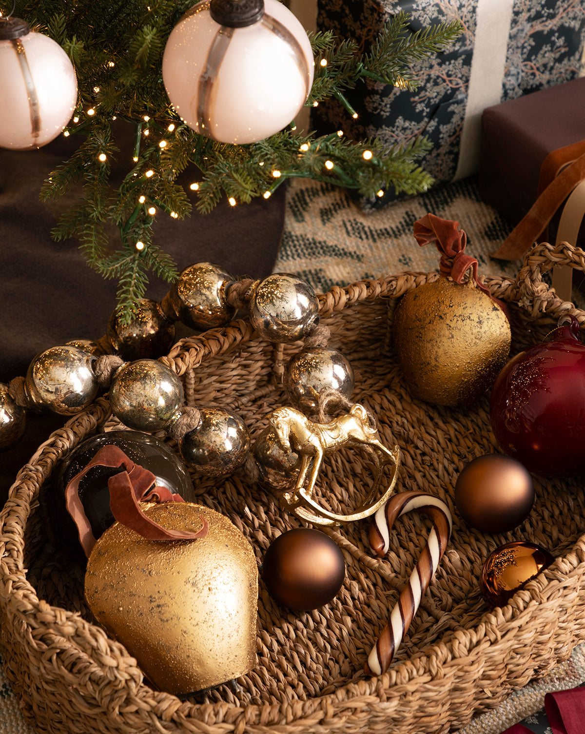 A woven basket with gold, silver, and red holiday décor sits under a decorated Christmas tree, accented by the Antique Gold Metal Bell with Velvet Ribbon for extra festive charm, by McGee & Co. Wrapped gifts are placed nearby.