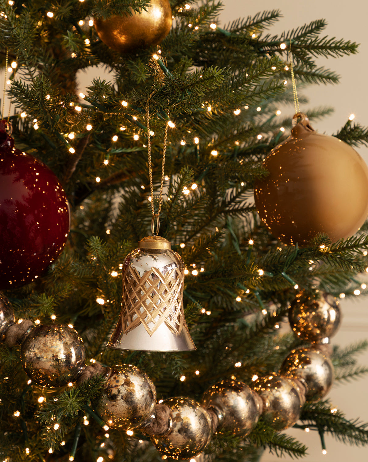A close-up of a decorated Christmas tree featuring gold and red ornaments, a Hand-Painted Mercury Glass Bell Ornament, warm white string lights, and a garland of shiny silver beads creates festive holiday décor, by McGee & Co.