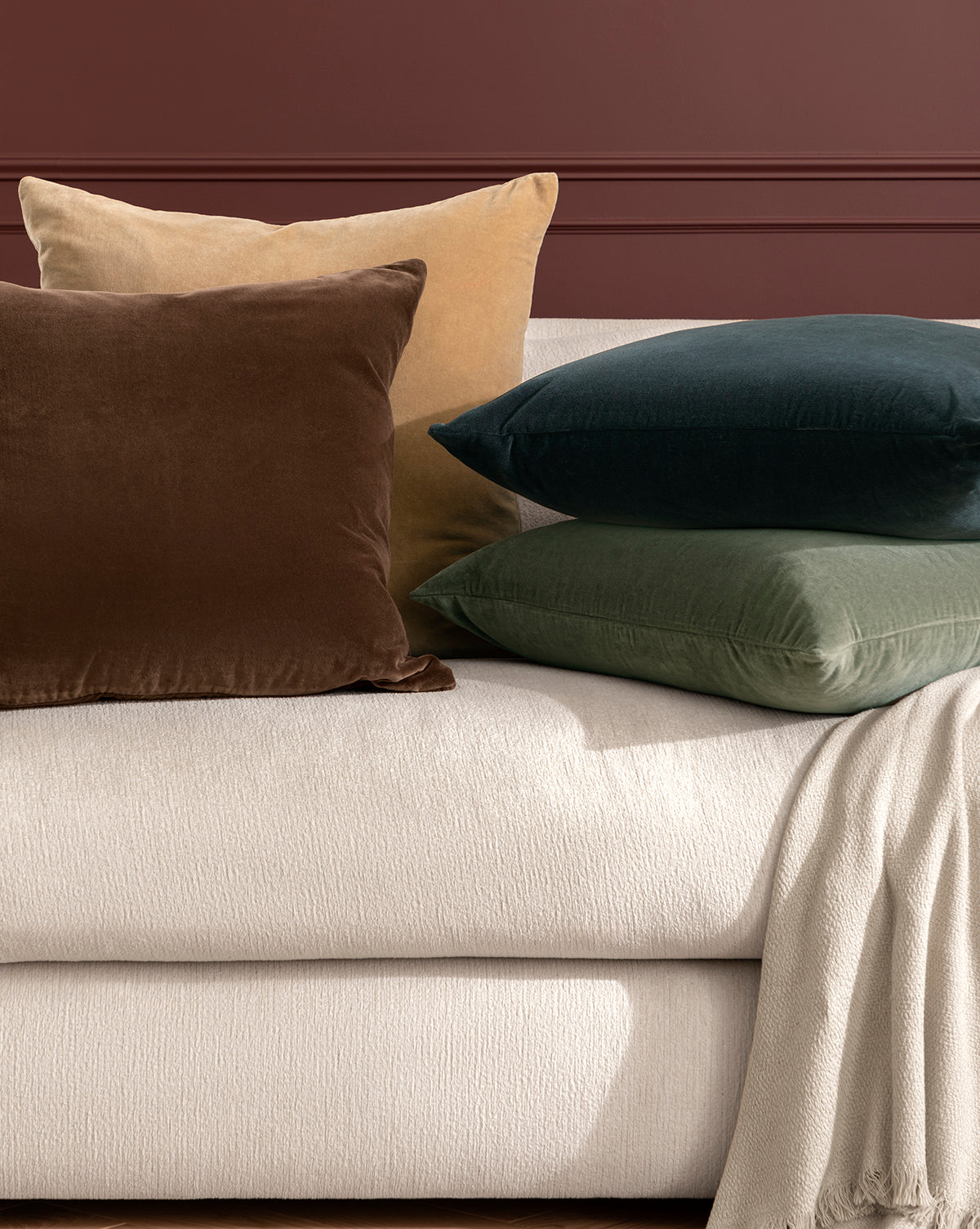 Four Bellevue Velvet Pillow Covers in brown, beige, dark green, and olive green add elegance to a light upholstered sofa with a cream blanket, set against a dark reddish-brown wall, by McGee & Co.