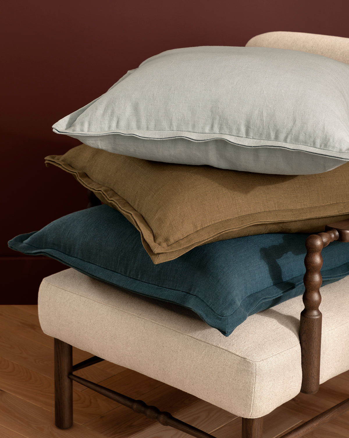Three large Liam Double Flange Pillow Covers in light gray, mustard yellow, and deep teal are stacked on a beige upholstered bench with a wooden leg and spindle arm, set against a burgundy wall and wood floor, by McGee & Co.