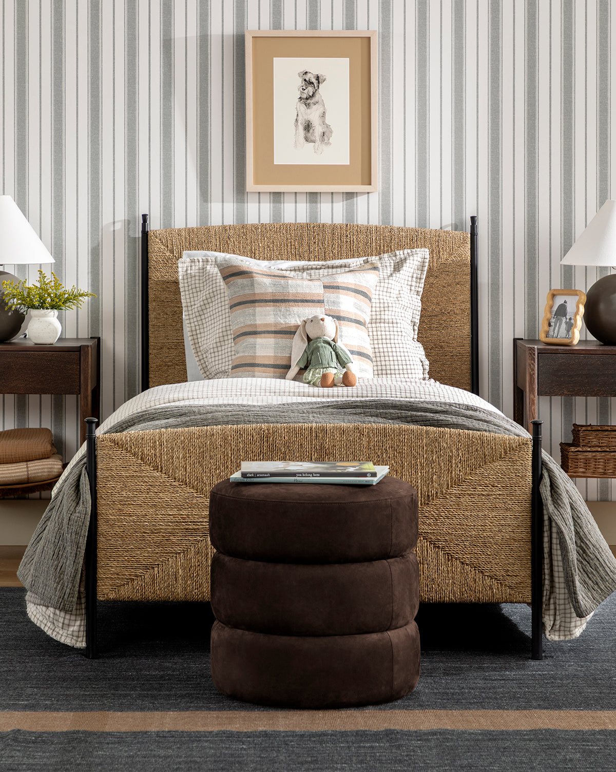 A cozy bedroom featuring a woven bed, plaid bedding, plush bunny toy, and brown ottoman. Nightstands with lamps and decor flank the bed; Hawes Stripe Wallpaper and a framed dog drawing decorate the wall behind, by McGee & Co.