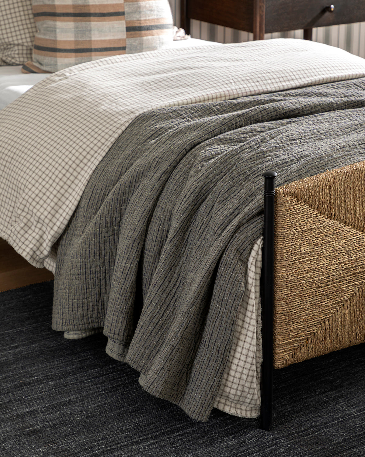A cozy bed with a checkered duvet, textured gray blanket, and plaid pillows features the charming Cecily Kids Quilt. A woven rattan footboard and a dark rug enhance the warm, inviting, rustic vibe by McGee & Co.