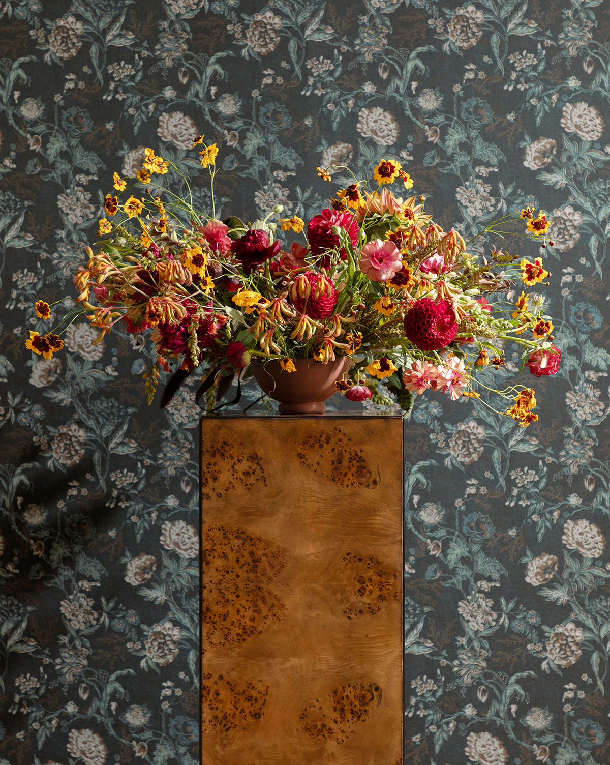 A vibrant bouquet in a brown vase sits on a wooden pedestal, set against vintage-inspired Hestercombe Floral Wallpaper featuring a dark floral design, by McGee & Co.