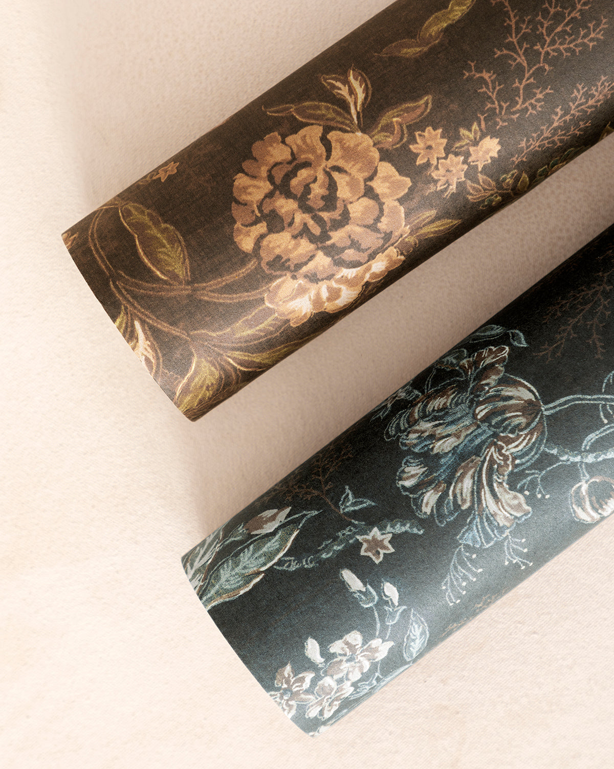 Two rolls of Hestercombe Floral Wallpaper—one with gold and brown flowers, the other with blue and green—are displayed on a light beige surface by McGee & Co.