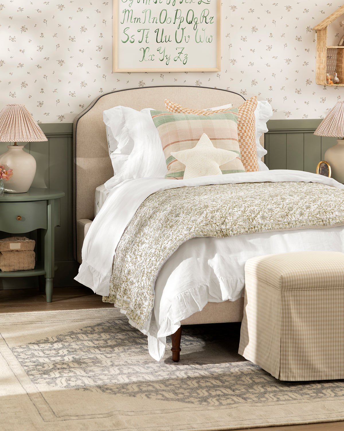 Cozy bedroom with a soft bed featuring the Seton Kids Quilt, white ruffled bedding, pastel pillows, a star cushion, green nightstand, matching lamps, floral wallpaper, and a decorative alphabet print above the headboard, by McGee & Co.