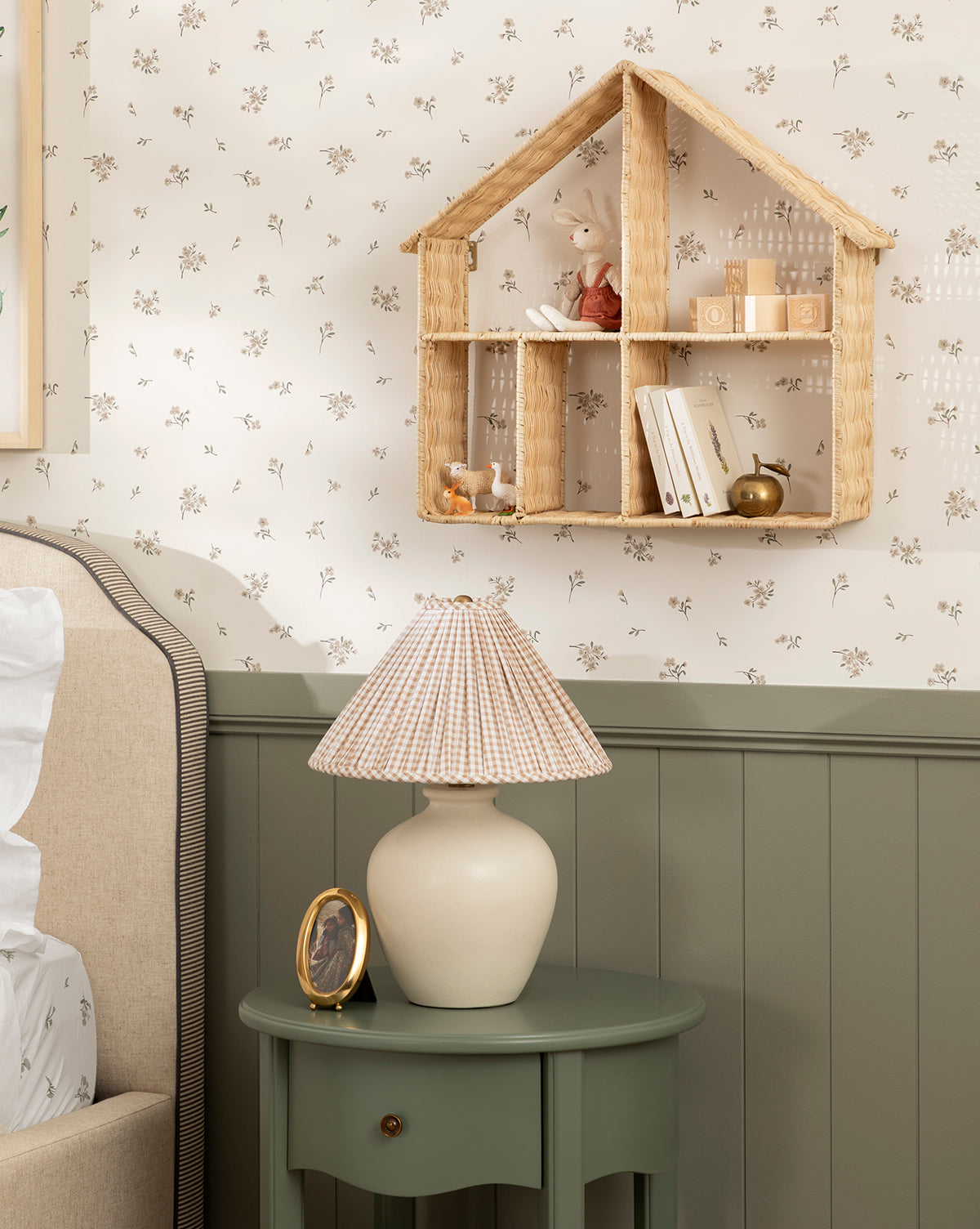 A cozy bedroom corner features a green nightstand with a lamp and photo, plus a woven house-shaped wall shelf holding books, a doll, and ornaments—all set against Wembley Floral Wallpaper by McGee & Co.