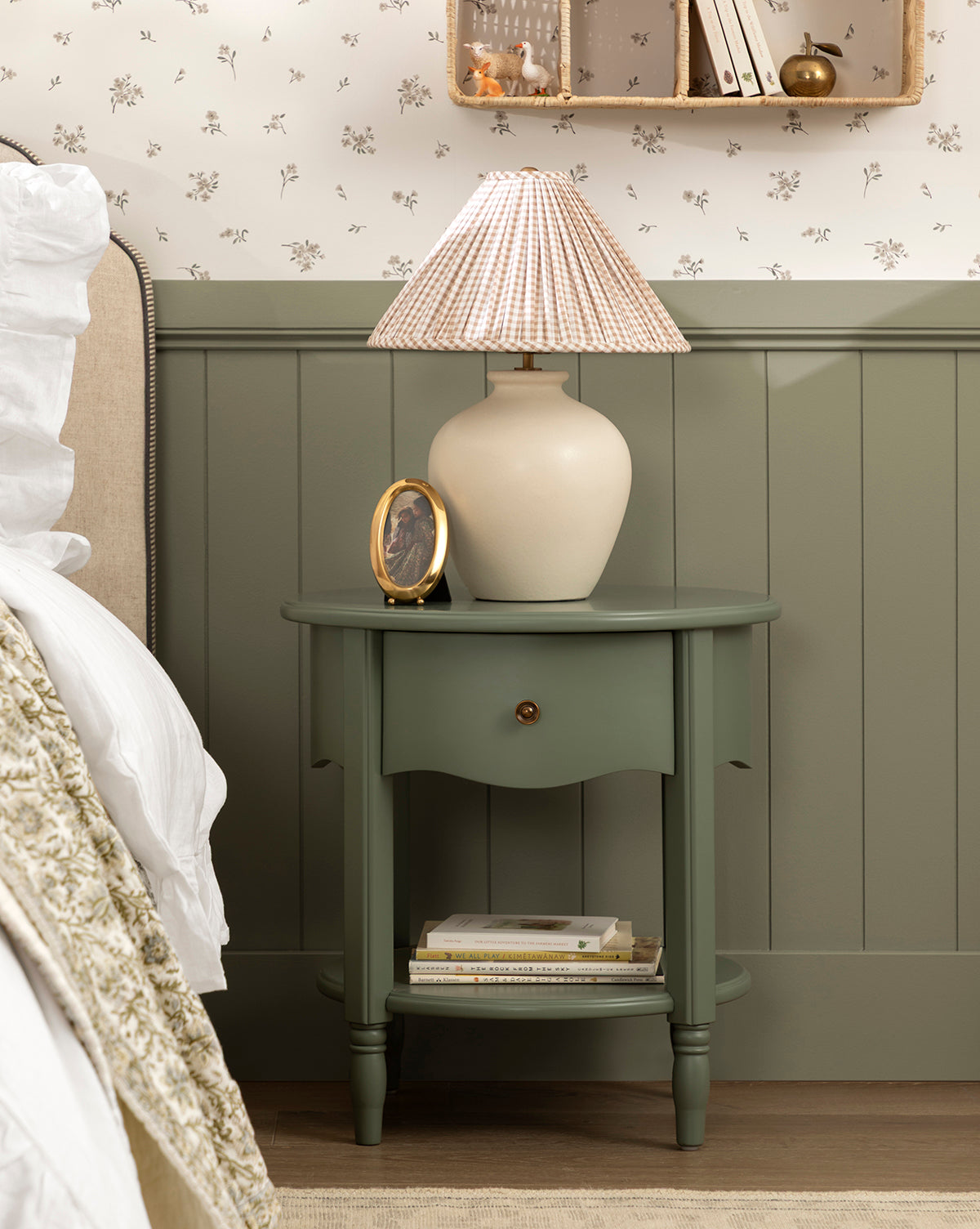 A green bedside table with a drawer and shelf holds the Bronwyn Table Lamp with a gingham pleated shade, a small oval photo frame, and several books; set beside a beige bed and floral wallpaper by McGee & Co.