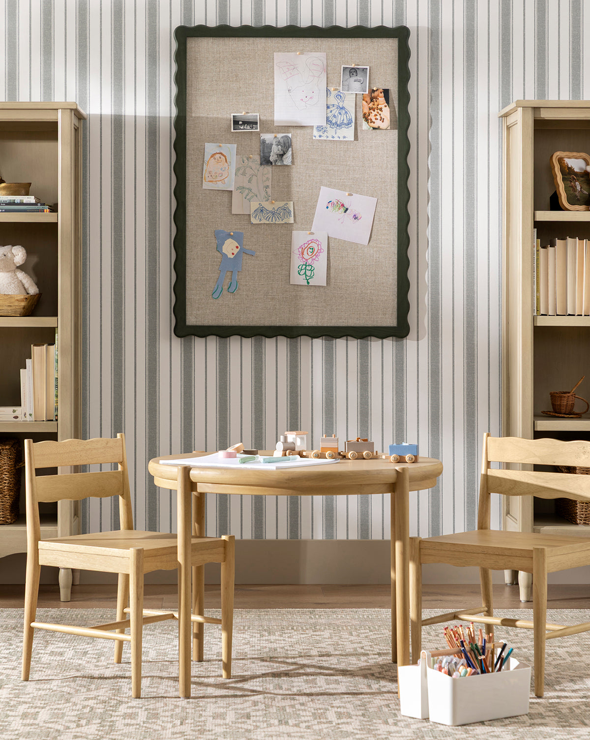 A children’s playroom with a small wooden table, two chairs, art supplies, toy cars, shelves of books and baskets, and a bulletin board with kids’ drawings—all set against playful Hawes Stripe Wallpaper with vertical stripes by McGee & Co.