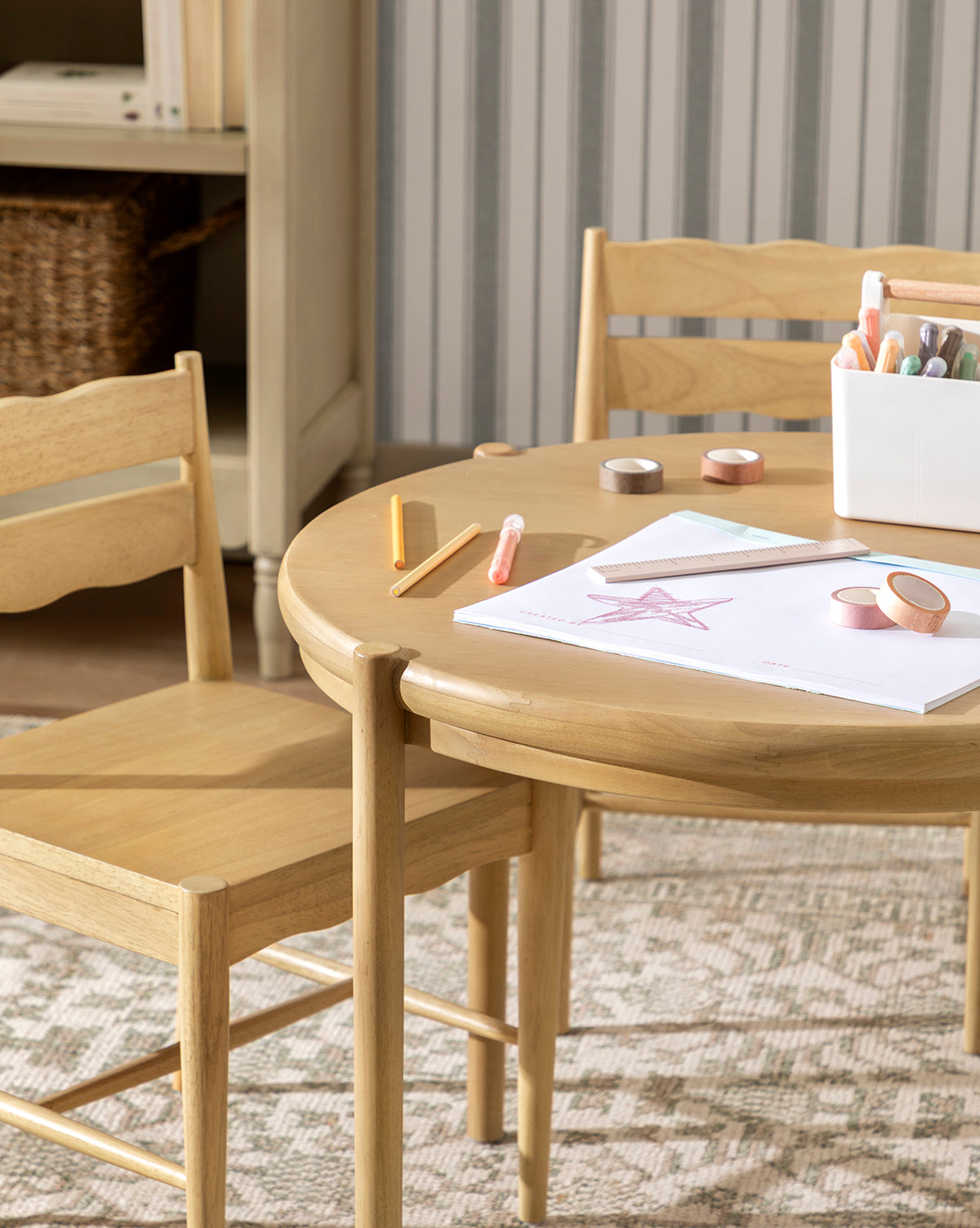 The Darcy Kids Table, paired with two chairs, displays art supplies, crayons, and markers. A star drawing and a white supply container rest on the table. Striped wallpaper and a woven basket are seen in the background, by McGee & Co.