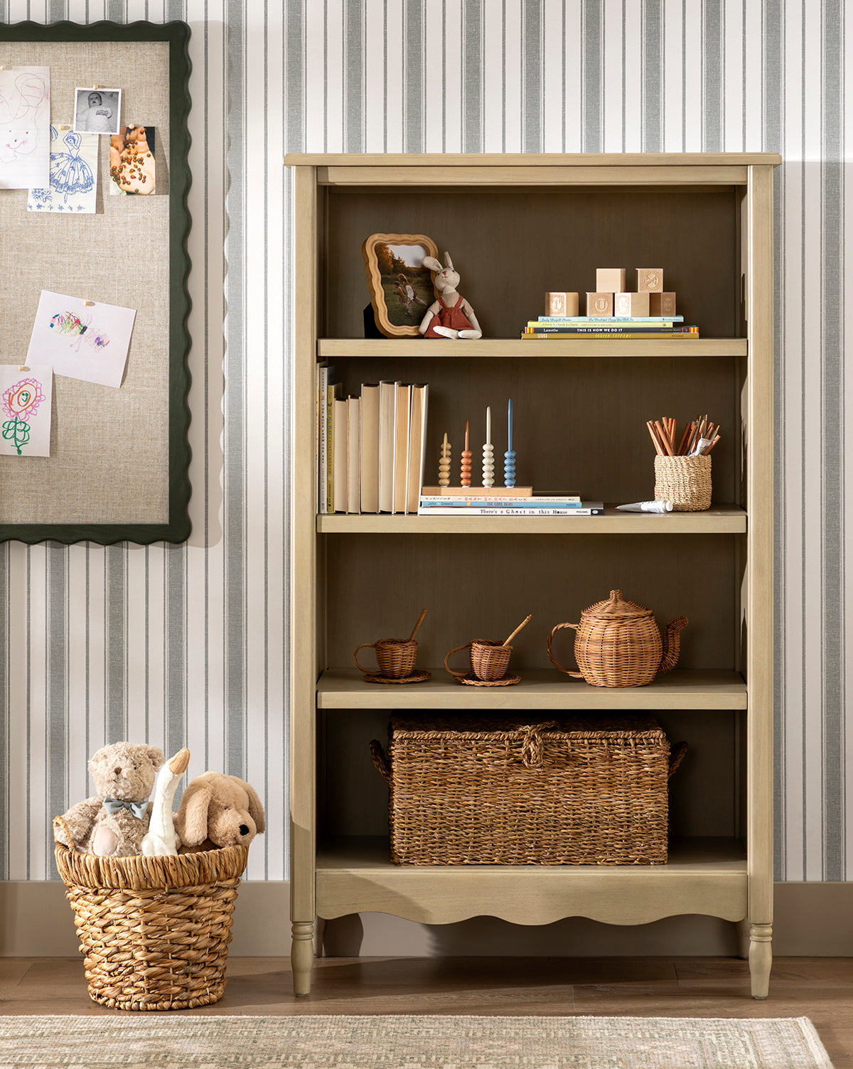 The Liberty Bookcase stands against a striped wall, displaying books, a toy rabbit, wooden stacking toys, cups, and baskets. Childrens drawings are pinned to a corkboard on the left, by McGee & Co.