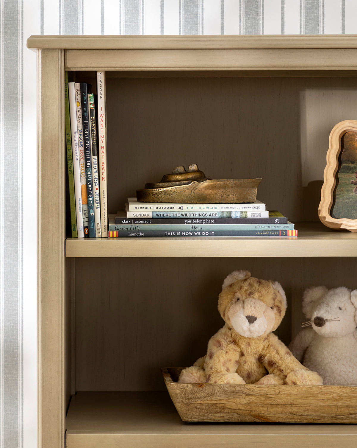 A wooden bookshelf holds childrens books on upper shelves and two stuffed animals—a lion and a white bear—on the lower shelf, with striped nautical wallpaper. A Cast Aluminum Boat Object adds a stylish maritime accent to the space, by McGee & Co.