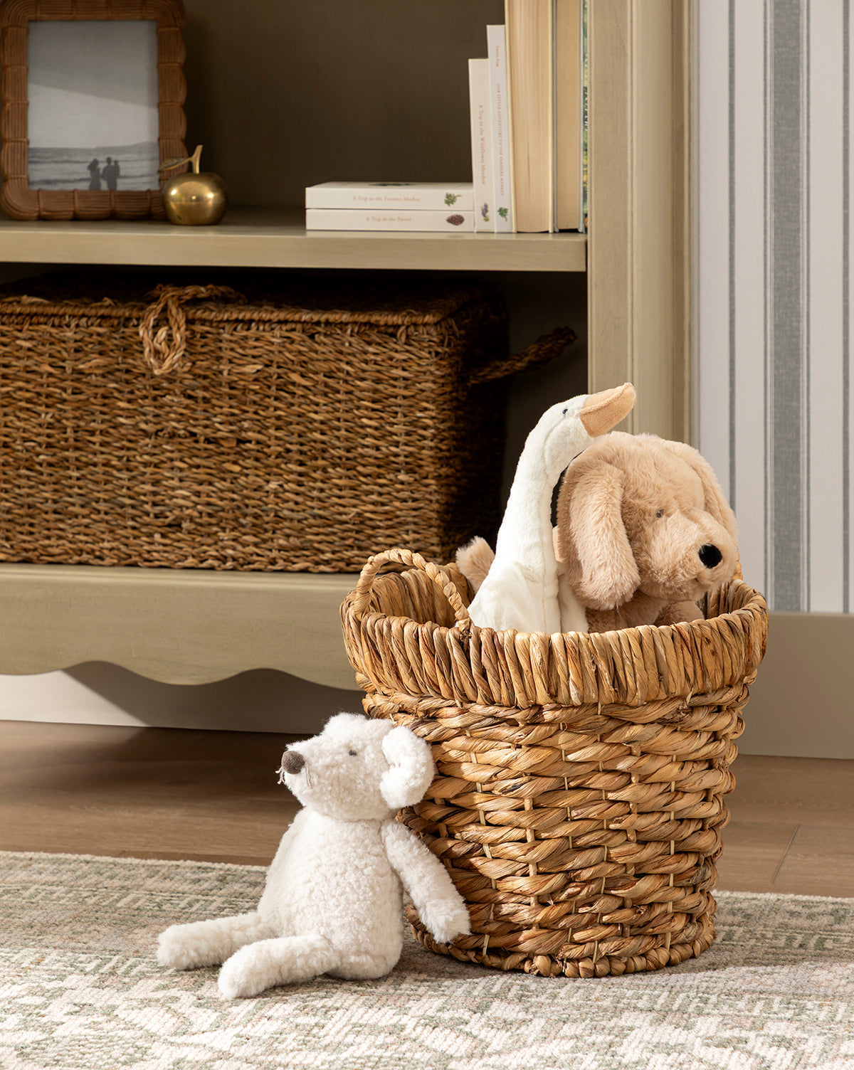 A decorative storage basket with stuffed animals—a dog and a duck—sits on a rug by a white teddy bear. Nearby, Water Hyacinth Baskets with Handles (Set of 2) and books add charm to the shelf with framed decor, by McGee & Co.