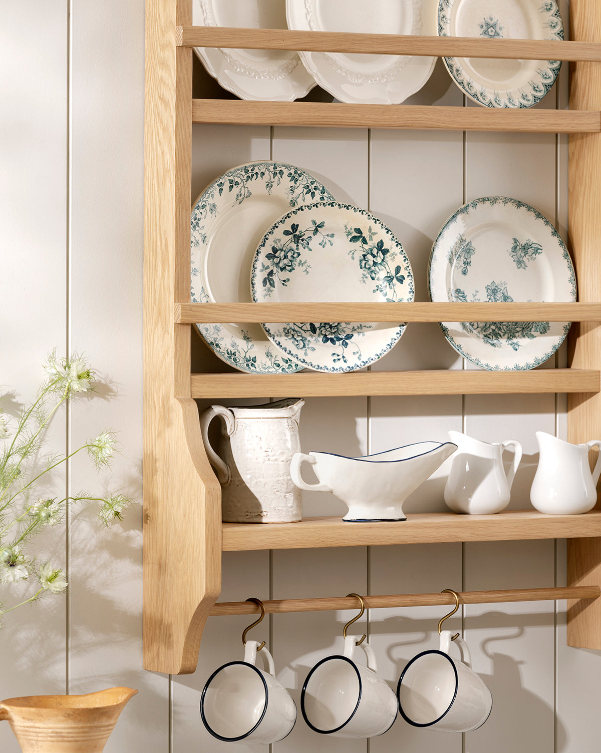 The Edna Kitchen Shelf displays floral-patterned plates, white pitchers, and teacups hanging from hooks. To the left, a light-toned wall and a vase of delicate greenery complete the inviting kitchen scene by McGee & Co.