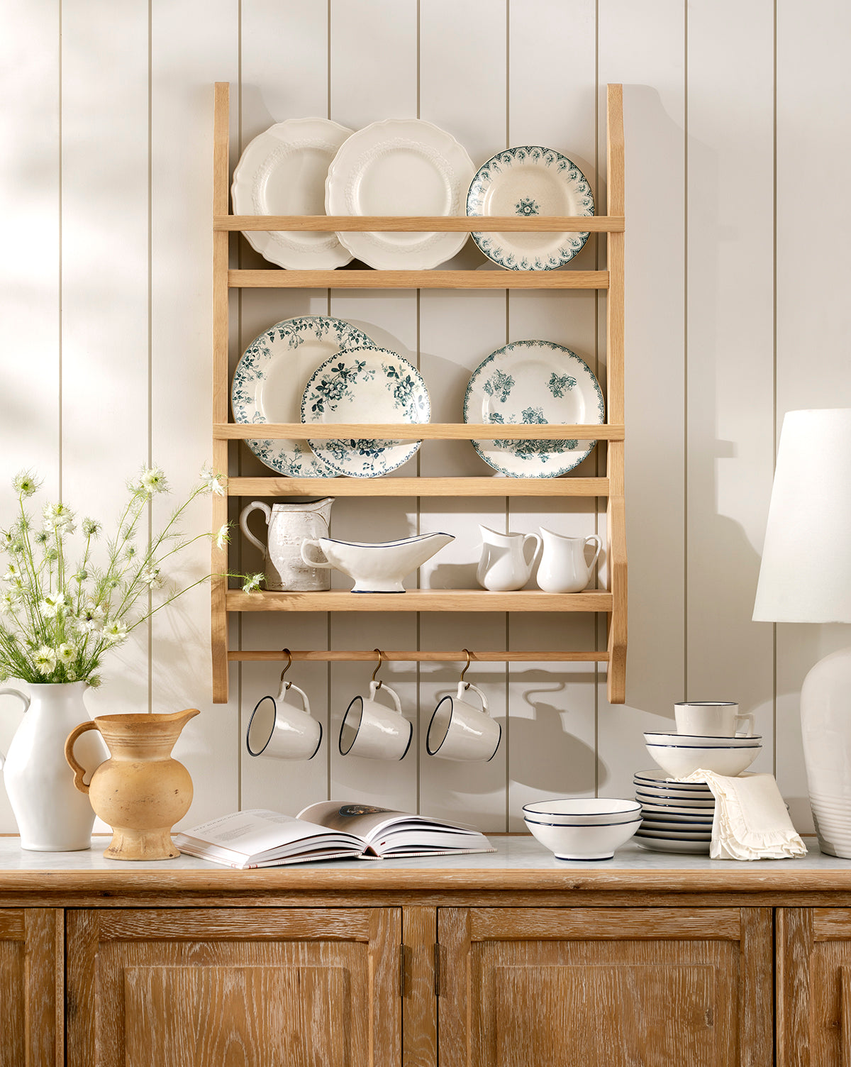 The Edna Kitchen Shelf on a white paneled wall displays white and floral plates, bowls, cups, and pitchers. Beneath it, a wooden counter holds an open book, stacked dishes, a vase with flowers, and a yellow jug by McGee & Co.