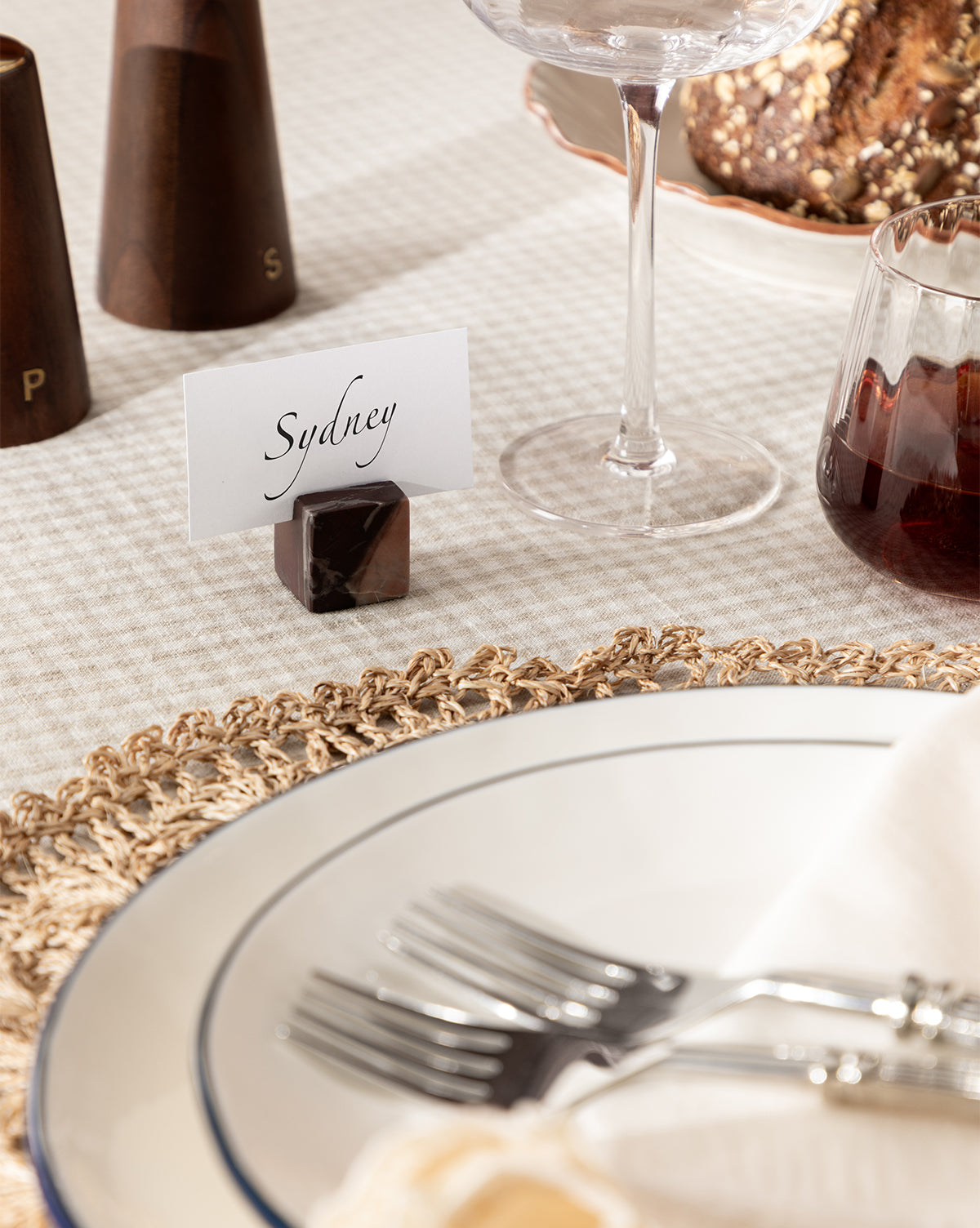 A close-up of an elegant holiday table features Auburn Marble Place Card Holders (Set of 4) with a card reading Sydney, forks on a white plate atop a woven placemat, and wine glasses nearby, by McGee & Co.