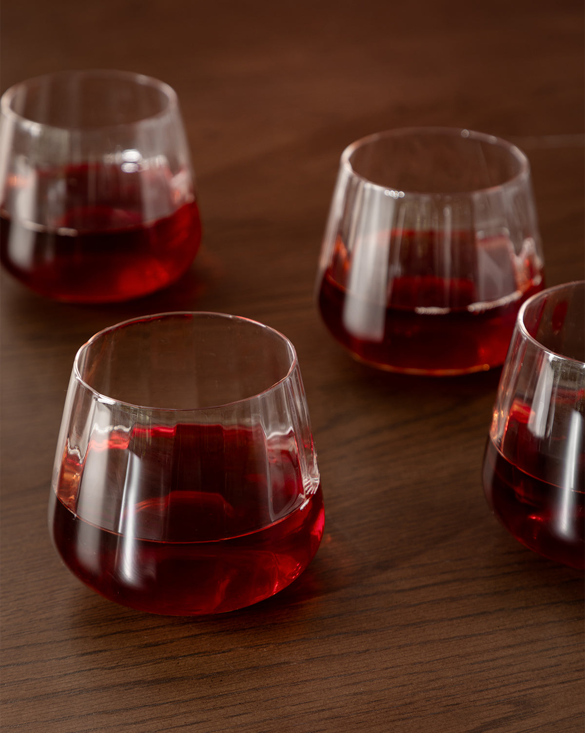 A set of four Langford Tumblers, filled with red liquid, rests on a wooden surface—their reflections shimmering and highlighting the elegance of this glassware set by McGee & Co.