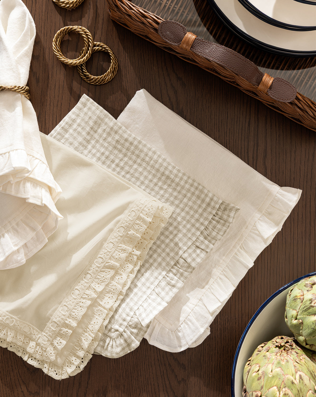 The Lace Trim Napkins (Set of 4), featuring beige and white vintage-inspired fabric with ruffled lace edges, are folded on a wooden table, paired with gold napkin rings, stacked plates in a wicker tray, and a bowl of artichokes by McGee & Co.