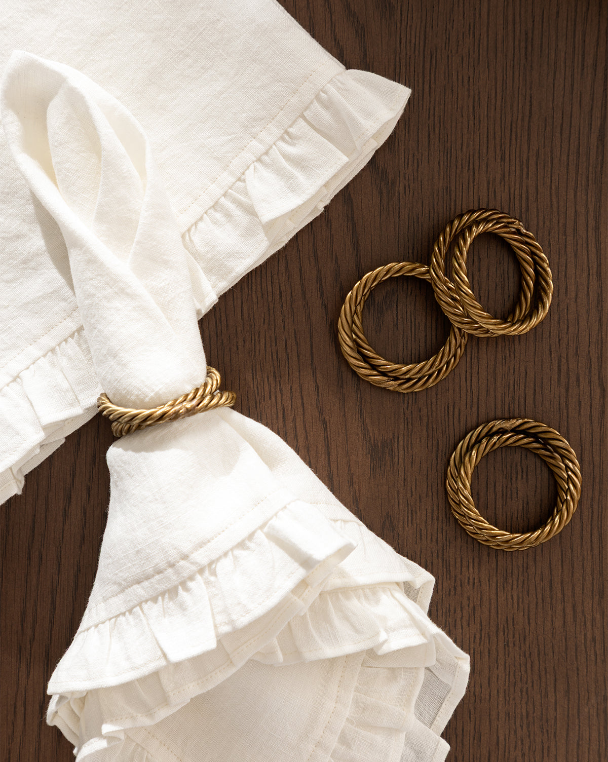 A folded white Catania Linen Napkin with ruffled edges is held by a gold twisted napkin ring, with three more gold napkin rings beside it on a wooden surface, by McGee & Co.