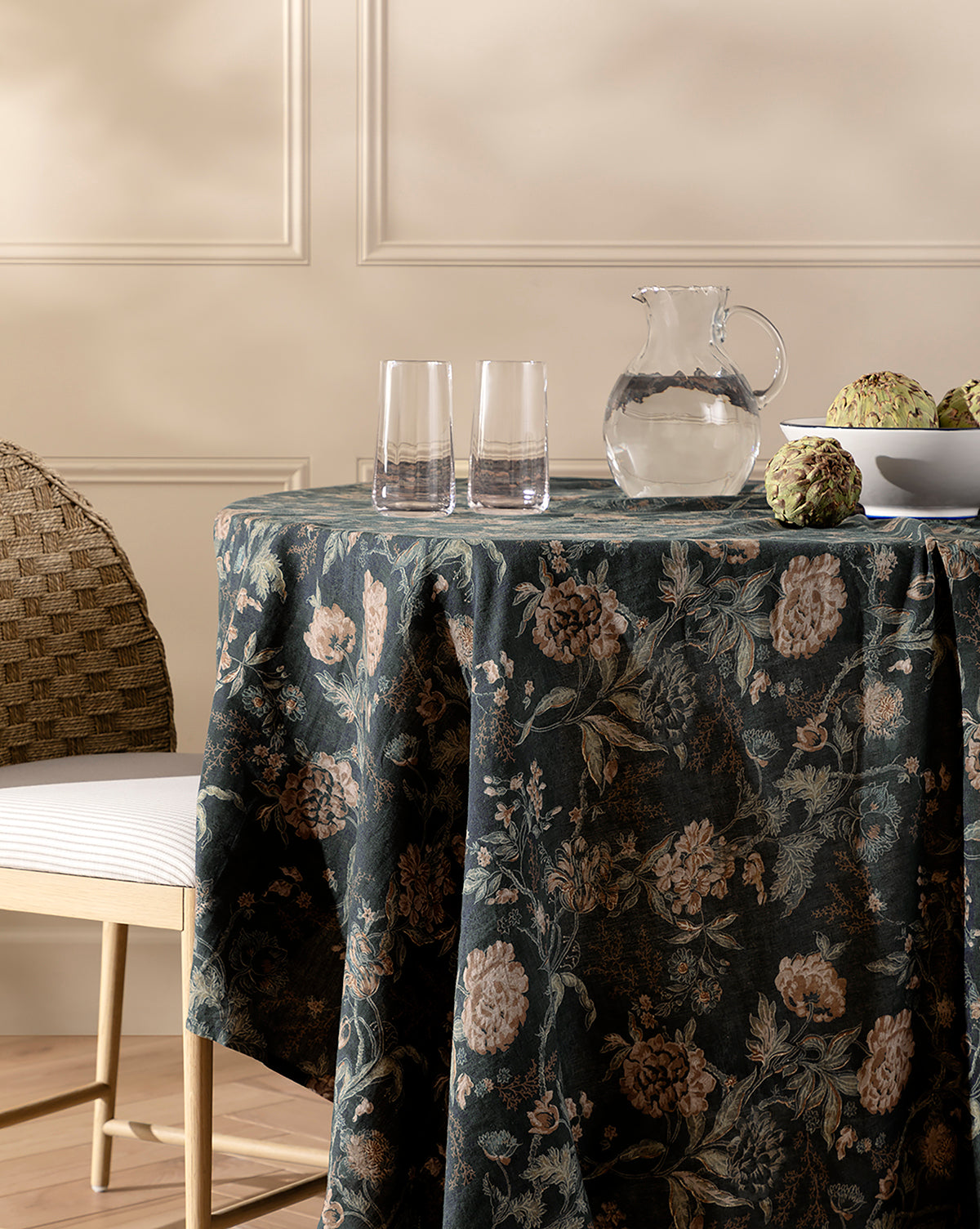 A round table draped with a dark Hestercombe Tablecloth holds a clear glass pitcher, two empty glasses, and a white bowl of artichokes. A woven chair with a white cushion sits beside the table against a beige wall with molding, by McGee & Co.
