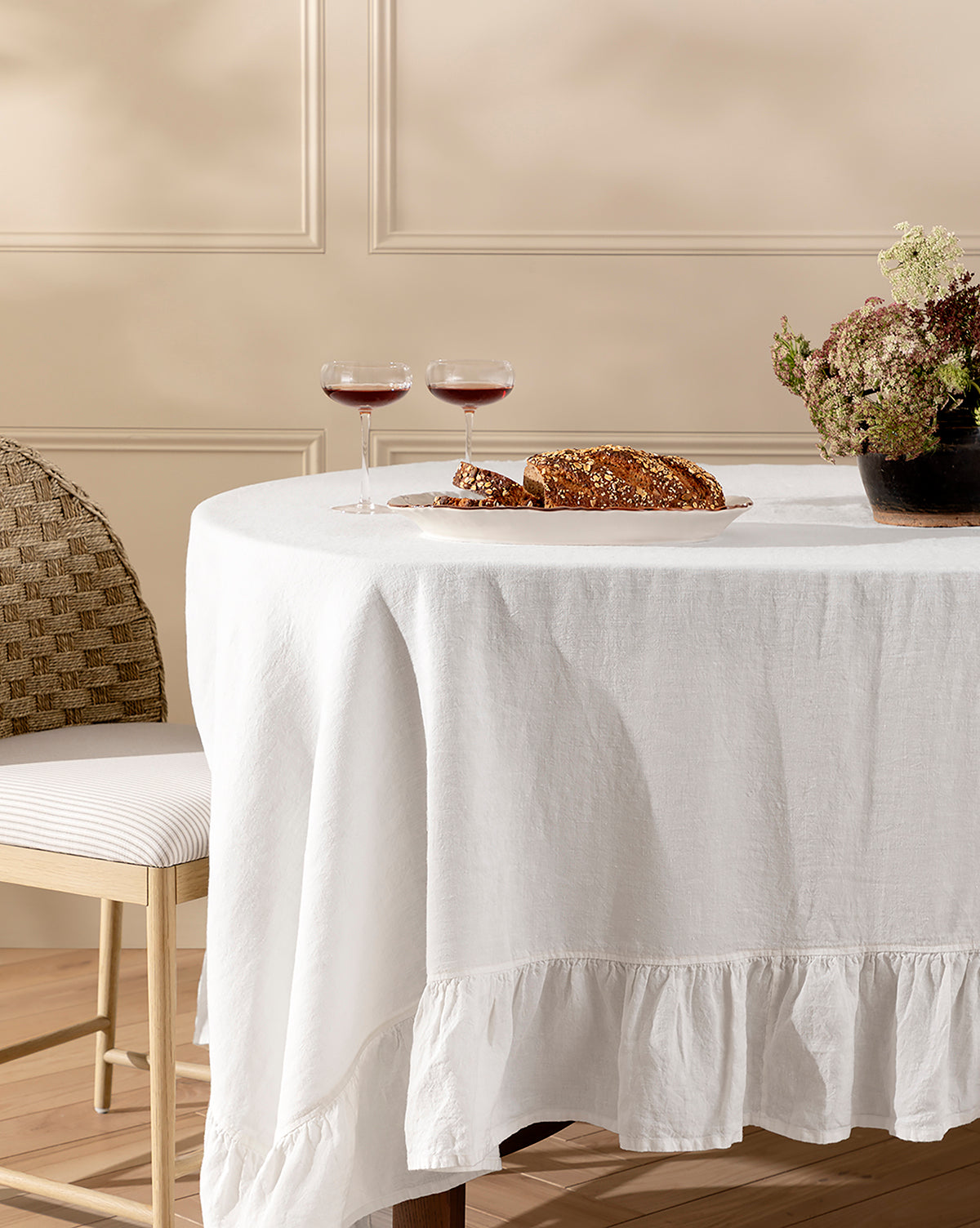 A round table draped with the Catania Linen Ruffle Tablecloth is set with two glasses of red wine, a loaf of bread on a cutting board, and a vase of flowers. A cushioned chair with a woven back sits nearby, by McGee & Co.