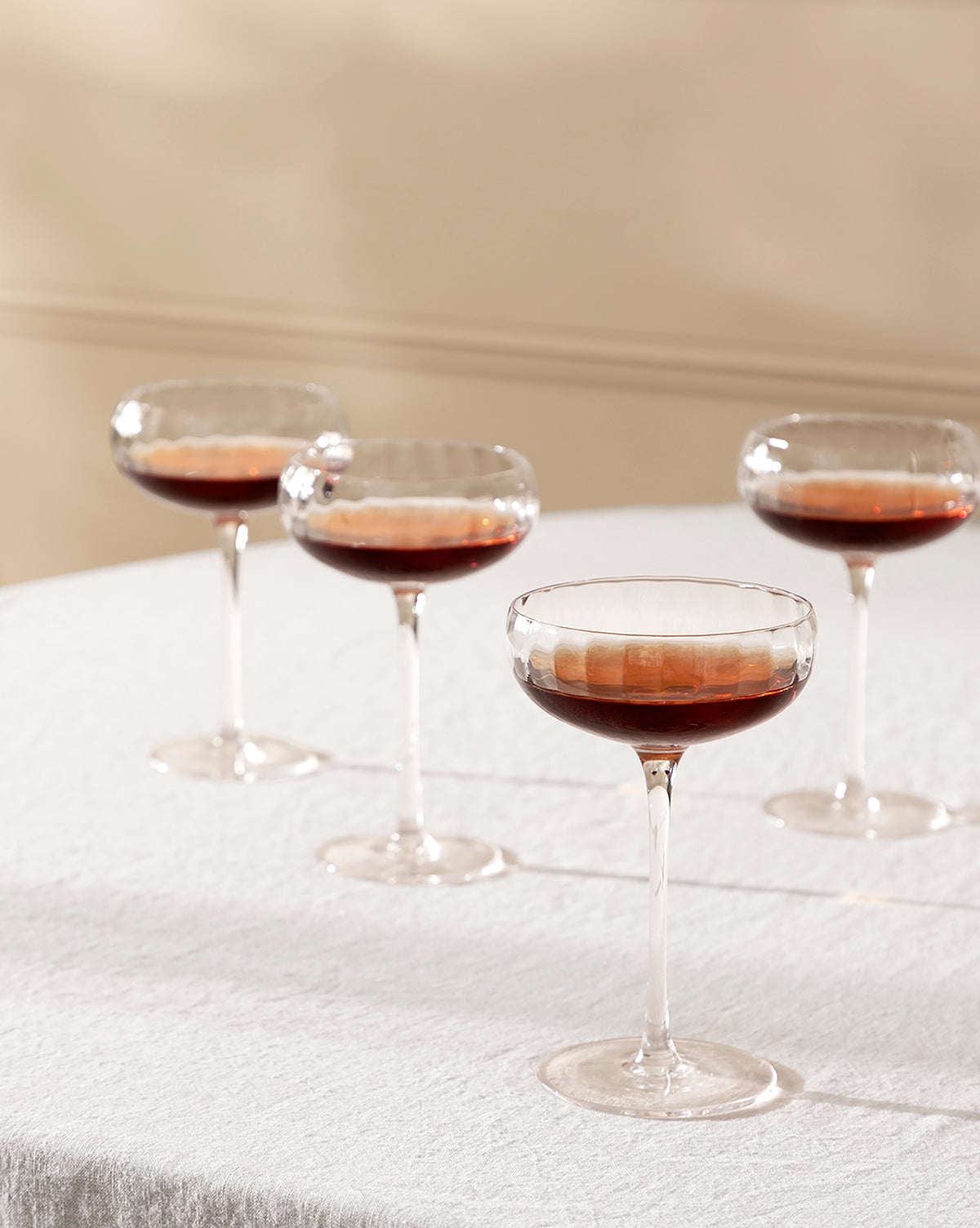 A set of four Langford Coupe glasses, each filled with a dark brown liquid, sits on a textured white tablecloth against a soft beige background—showcasing the elegant Langford Coupe (Set of 4), perfect for any occasion, by McGee & Co.