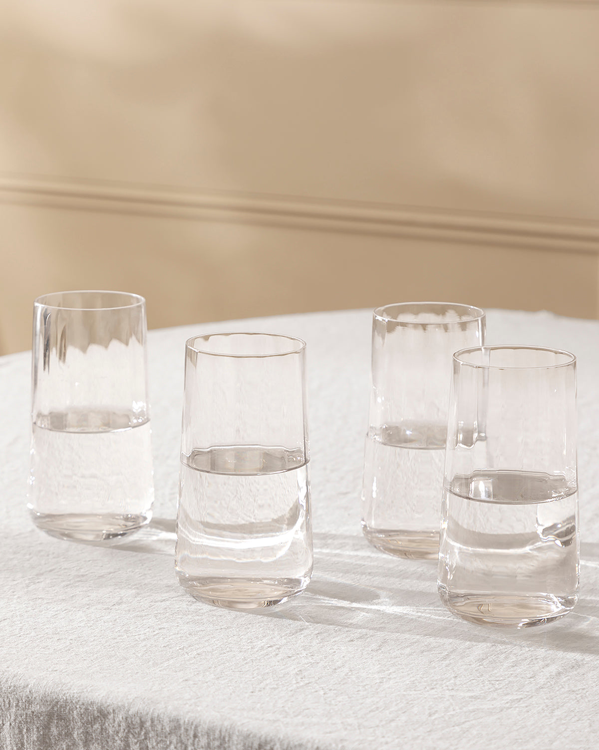 A set of four Langford Highball Glasses, each partially filled with water, is arranged on a white textured tablecloth against a soft beige background, by McGee & Co.