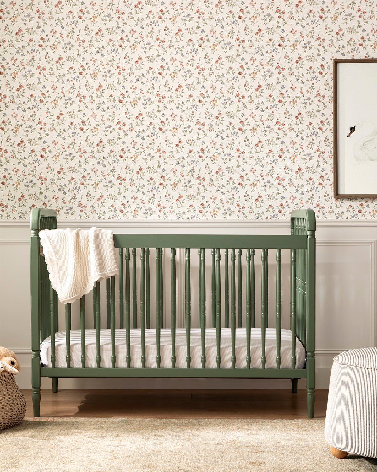 The Liberty 3-in-1 Convertible Spindle Crib w/Toddler Bed Conversion Kit in green, made from solid pine wood, stands before floral wallpaper with a white blanket draped over the side, by McGee & Co. A woven basket with a plush toy is nearby.