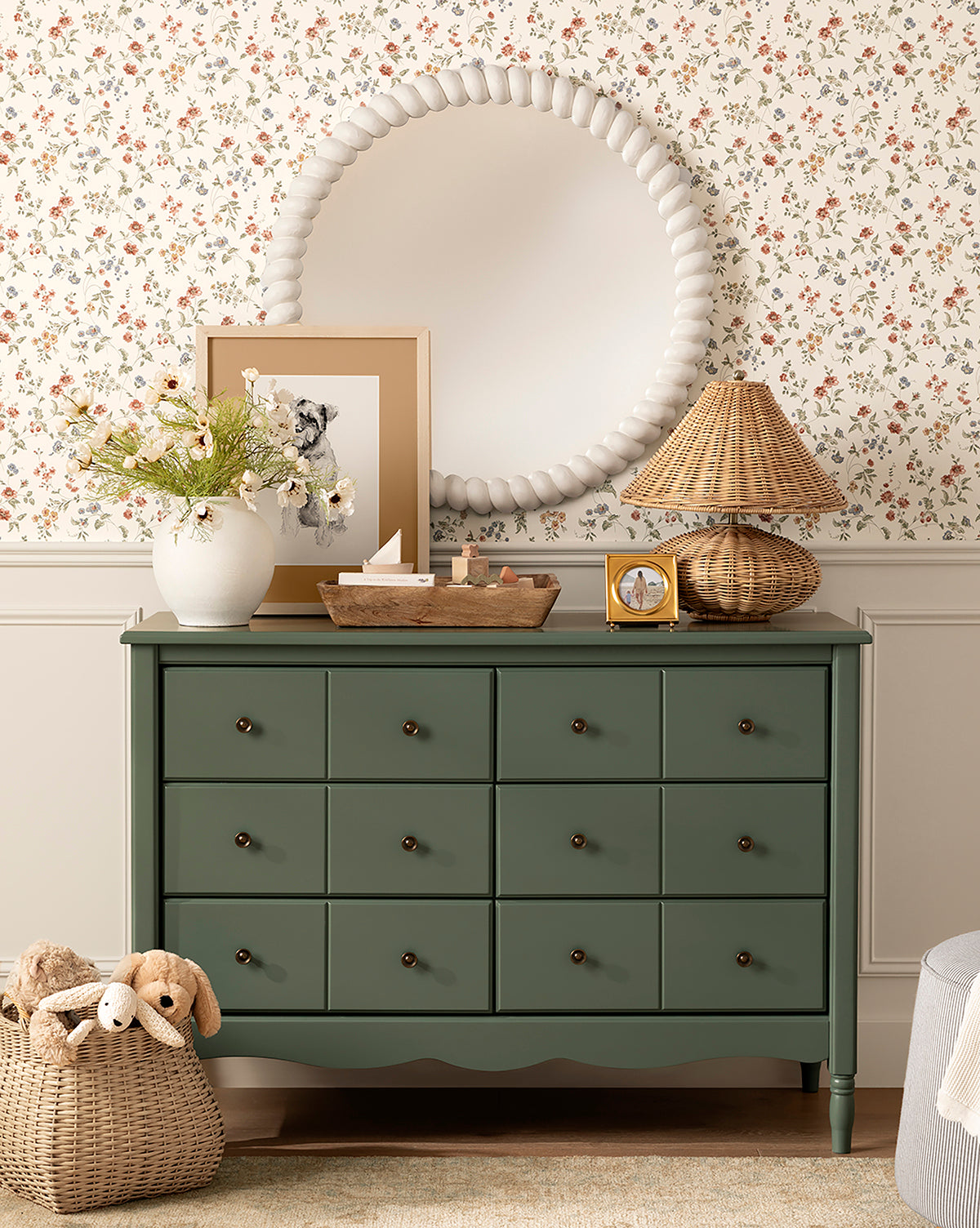 The Liberty 6-Drawer Assembled Dresser, made from sustainable pine with brass knobs, stands against floral wallpaper and is styled with a beaded mirror, framed art, wicker lamp, vase of flowers, wooden tray, and a basket of stuffed animals on the floor, by McGee & Co.