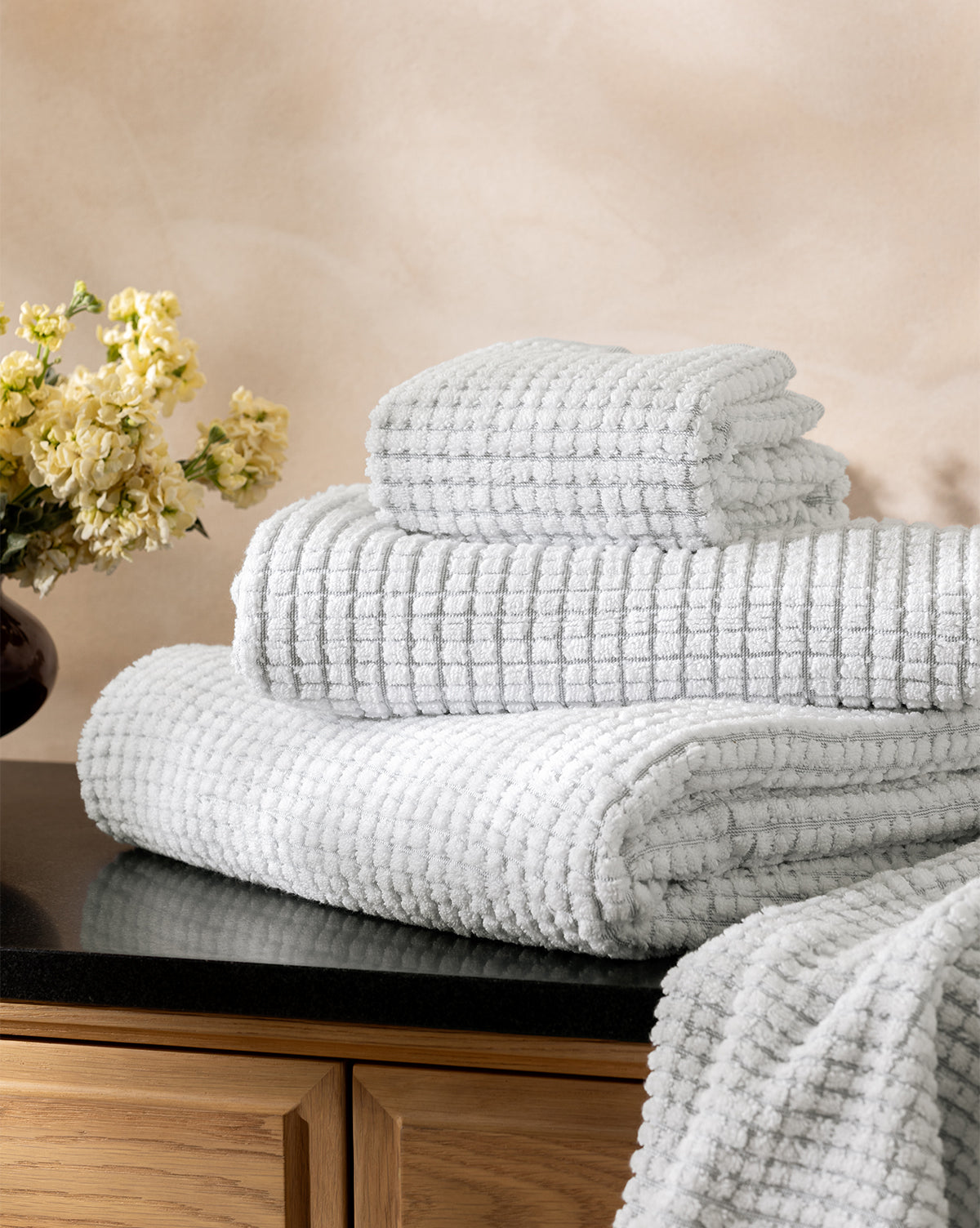 A neatly stacked set of Atrani Gray Organic Bath Collection waffle weave towels sits on a black countertop next to a vase of yellow flowers, against a light beige wall, by McGee & Co.