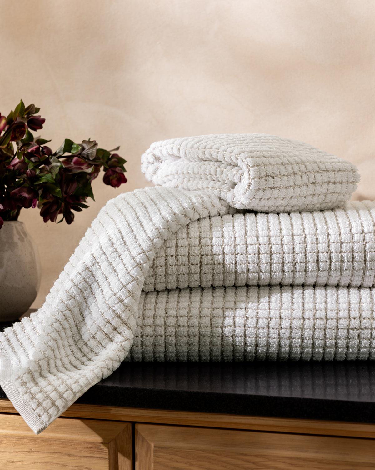 The Atrani Tan Organic Bath Collection, made of organic cotton with a waffle weave, is neatly folded on a dark countertop beside dark red flowers, set against light walls and wooden cabinets, by McGee & Co.