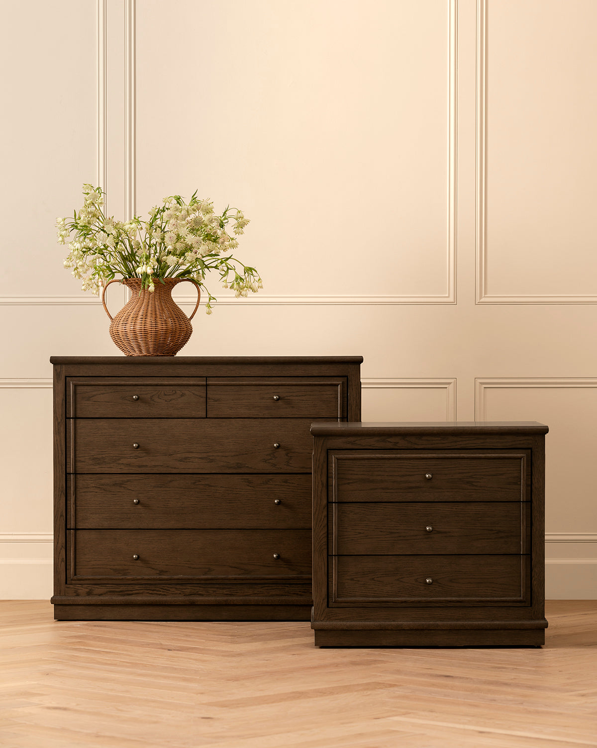 Two Yorkshire Nightstands with antique brass pulls, one taller and one shorter, stand side by side on a light herringbone floor. The taller nightstand holds a brown vase with white flowers against a cream-colored paneled wall, by McGee & Co.