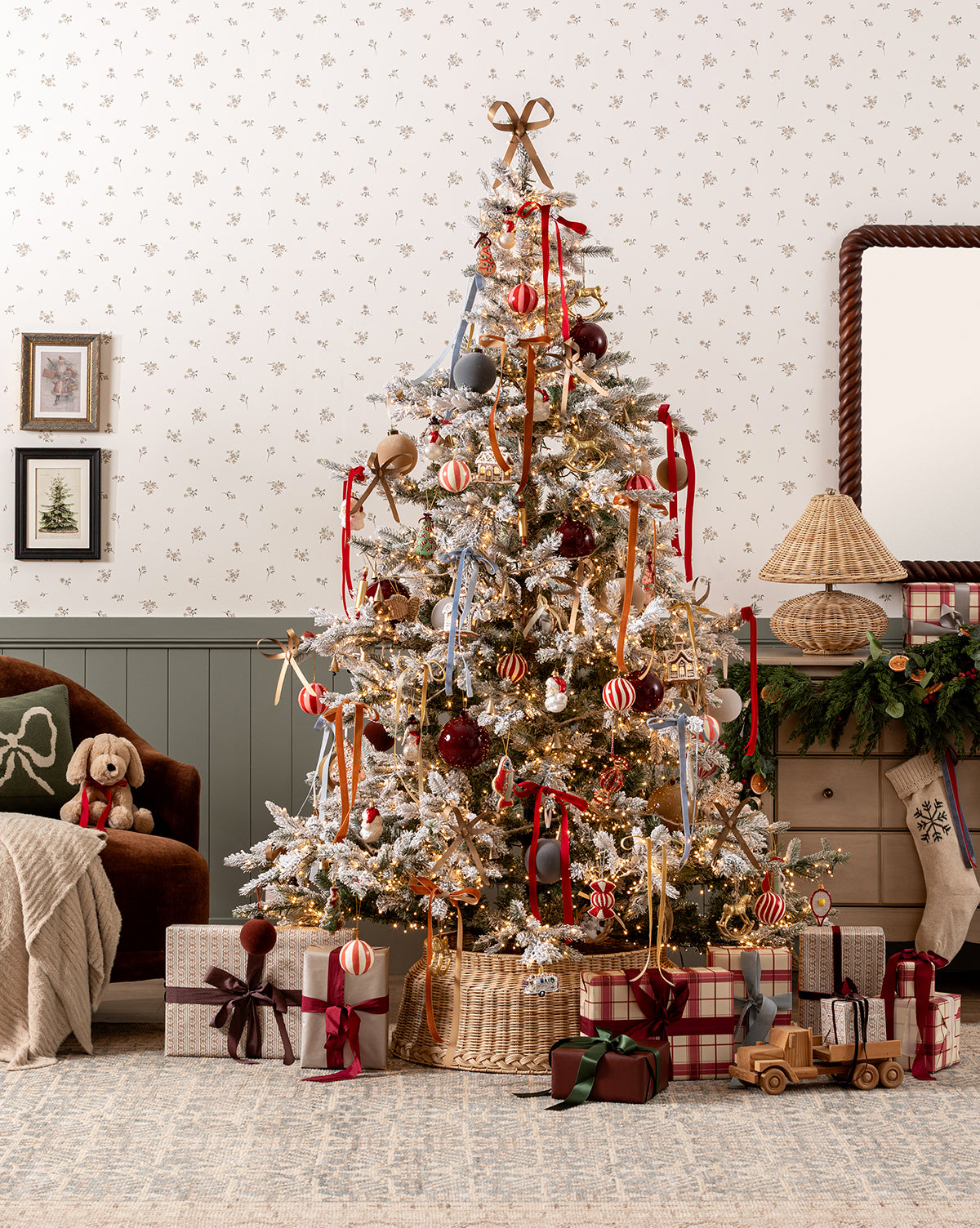 The Magical Wonderland Tree shines in a cozy living room, adorned with red and gold ornaments, ribbons, and lights. Gifts sit below the tree, surrounded by a plush dog toy, vintage wooden toys, gingerbread ornaments, and festive décor, by McGee & Co.
