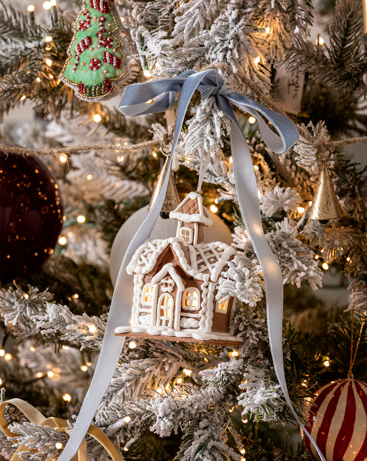 A snowy gingerbread house ornament with white icing and a blue ribbon hangs on the Magical Wonderland Tree, surrounded by lights, a green cookie ornament, and festive holiday decorations, by McGee & Co.