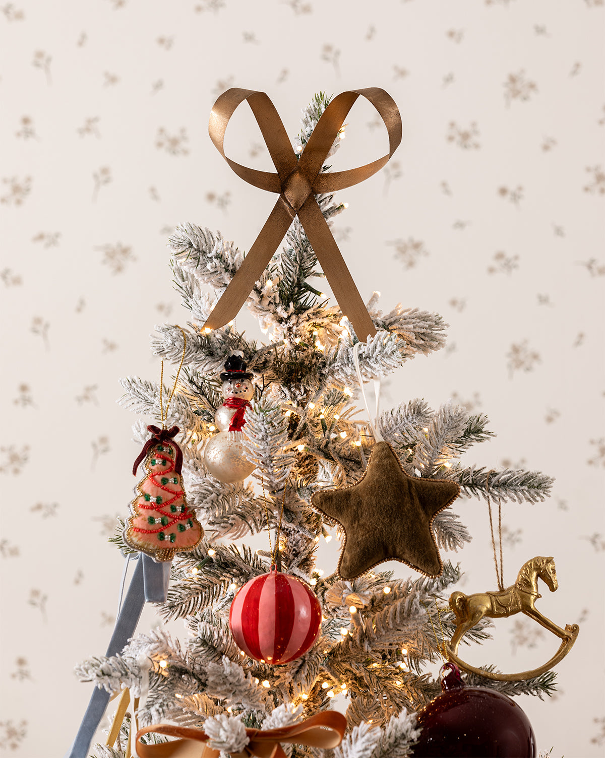 A small, snow-dusted holiday tree is decorated with festive style, featuring a gold ribbon topper, a Plush Beaded Star Ornament, a red-and-white striped ball, a rocking horse, a snowman, and warm white lights, by McGee & Co.