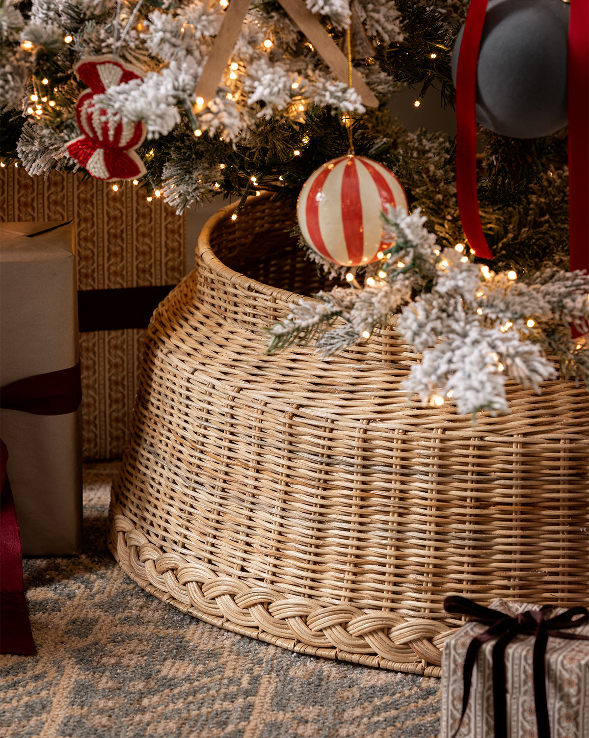 A Magical Wonderland Tree adorned with ornaments and lights stands in a woven tree collar. Wrapped gifts with ribbons rest nearby on a patterned rug, completing the charming holiday decor, by McGee & Co.