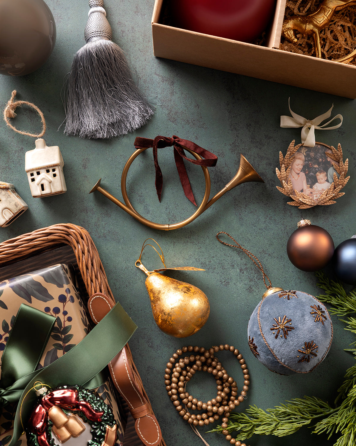 A flat lay of Christmas ornaments, featuring a gold horn with red ribbon, Laurel Picture Frame Ornament for a sentimental touch, golden pear, blue velvet ball, festive beads, ceramic house, wrapped gifts, tassel, and evergreen branches by McGee & Co.