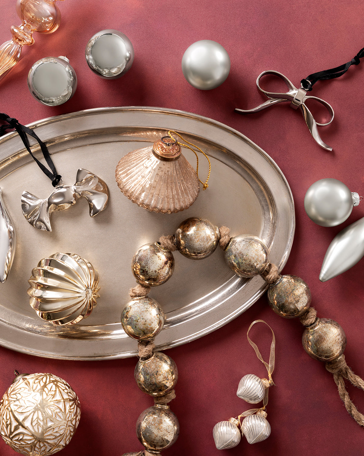 A silver tray with metallic ornaments, 3" Gold Baubles, glass baubles, decorative beads, and ribbon bows is arranged on a reddish background for an elegant holiday display with silver, gold, and pearl accents, by McGee & Co.