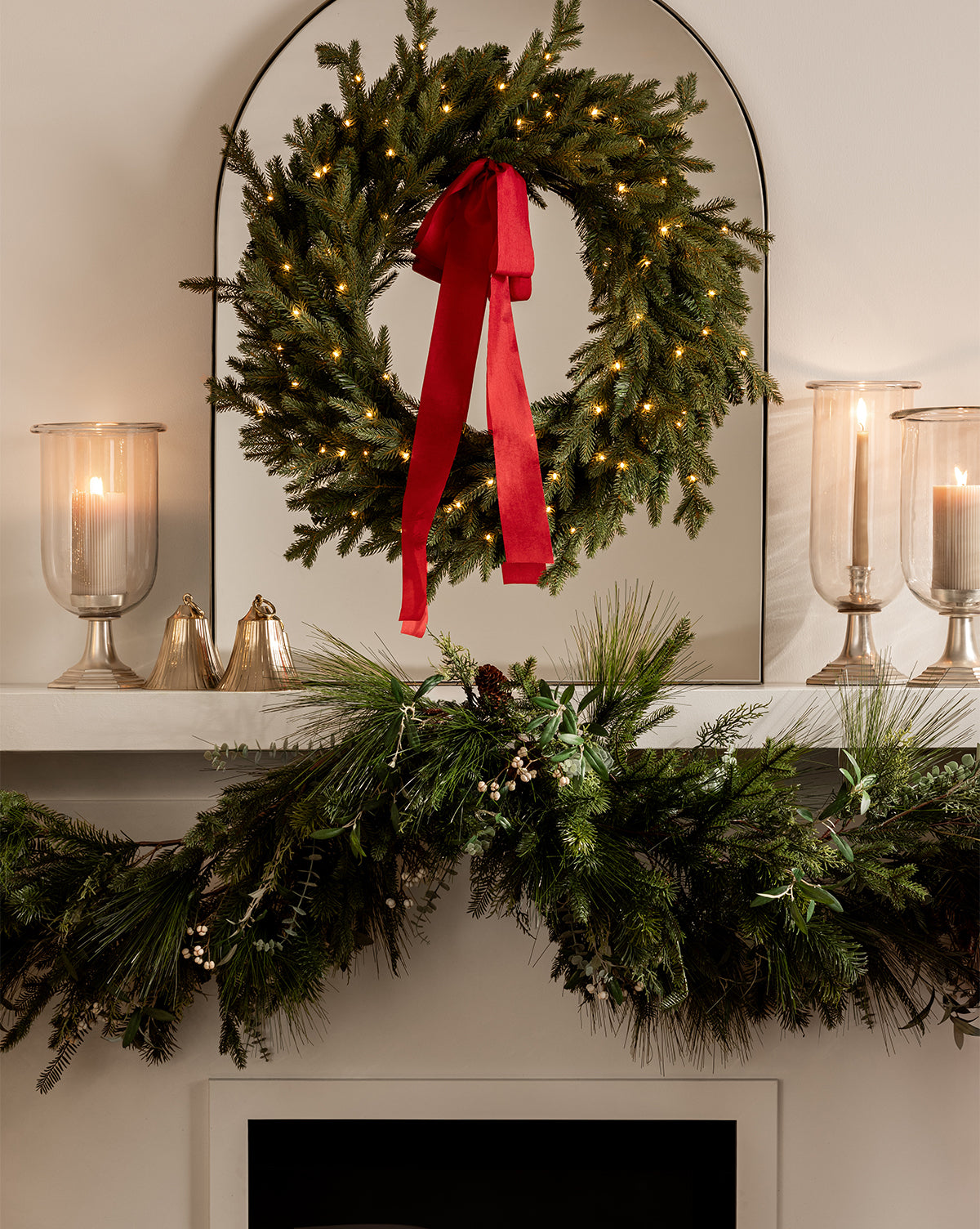 A festive mantel is adorned with a vintage-inspired green wreath with a red ribbon, two lit candles in glass holders, Polished Brass Bell ornaments, and a pine garland for charming holiday décor, by McGee & Co.