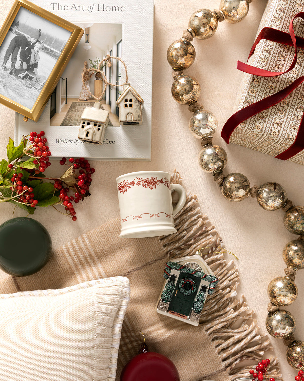 A cozy holiday scene with a beige blanket, book, mug, pillow, berry sprigs, silver garland, framed photo, two ornaments including the Festive Front Door Ornament, a wrapped gift with red ribbon, and house-shaped Christmas tree decorations, by McGee & Co.