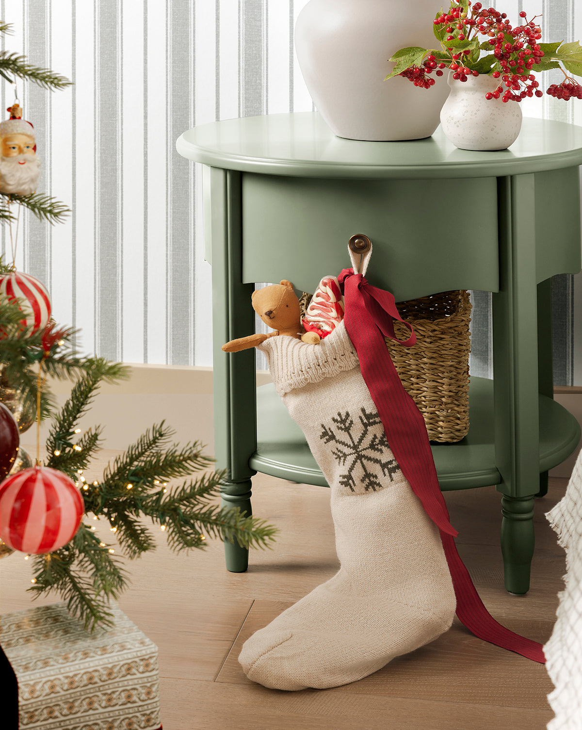 A cream-colored Bella Scalloped Stocking with Snowflake hangs on a green side table, filled with gifts and a reindeer plush, surrounded by festive holiday mantel decor, by McGee & Co.